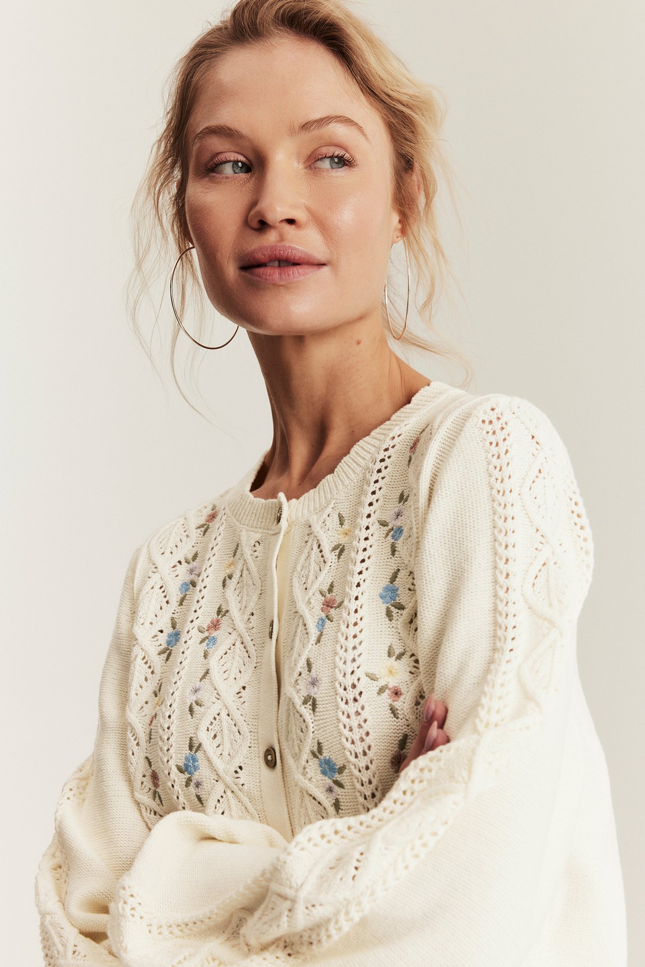 Front view of a woman wearing an off-white knitted cardigan with floral embroidery and large silver hoop earrings.