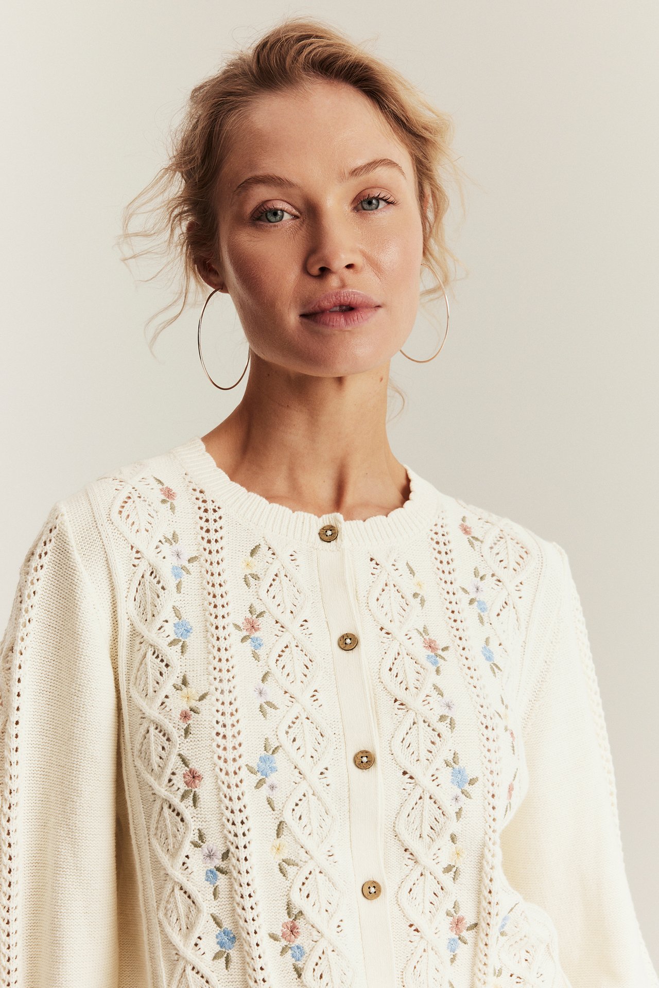 Front view: woman in off-white floral embroidered cotton cardigan with buttons, wearing silver hoop earrings.
