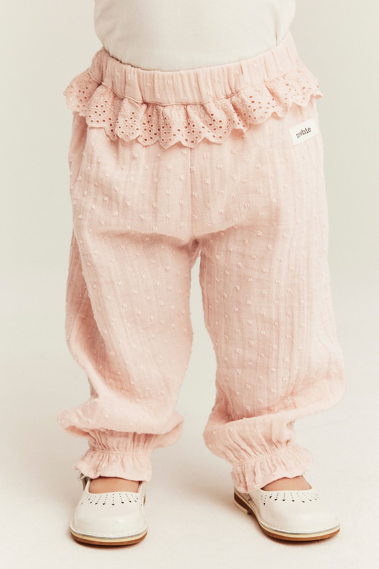 Vorderansicht: Babys Beine in hellrosa strukturierten Baumwollhosen mit Spitzenrüsche am Bund und weißen Schuhen.