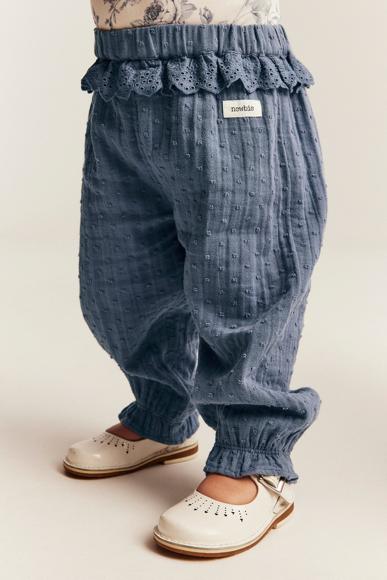 Nahaufnahme von Baby in blauer strukturierter Hose mit Spitzenrüsche, Gummizugbund und weißen Schuhen.