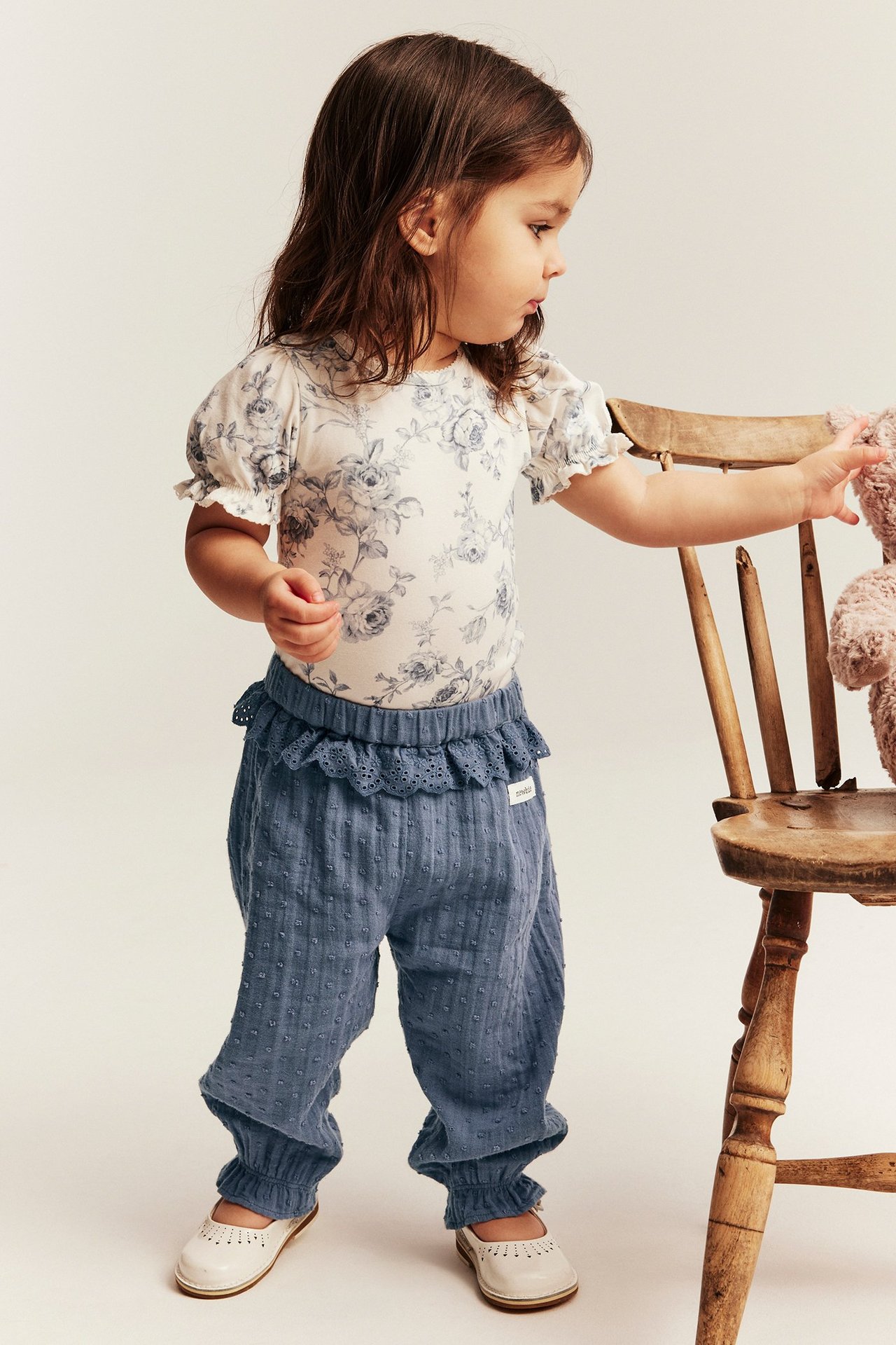 Vorderansicht: Kind trägt blaue Baumwollhose mit Punktstruktur und Spitzenrüschen, geblümtes Oberteil, weiße Schuhe.