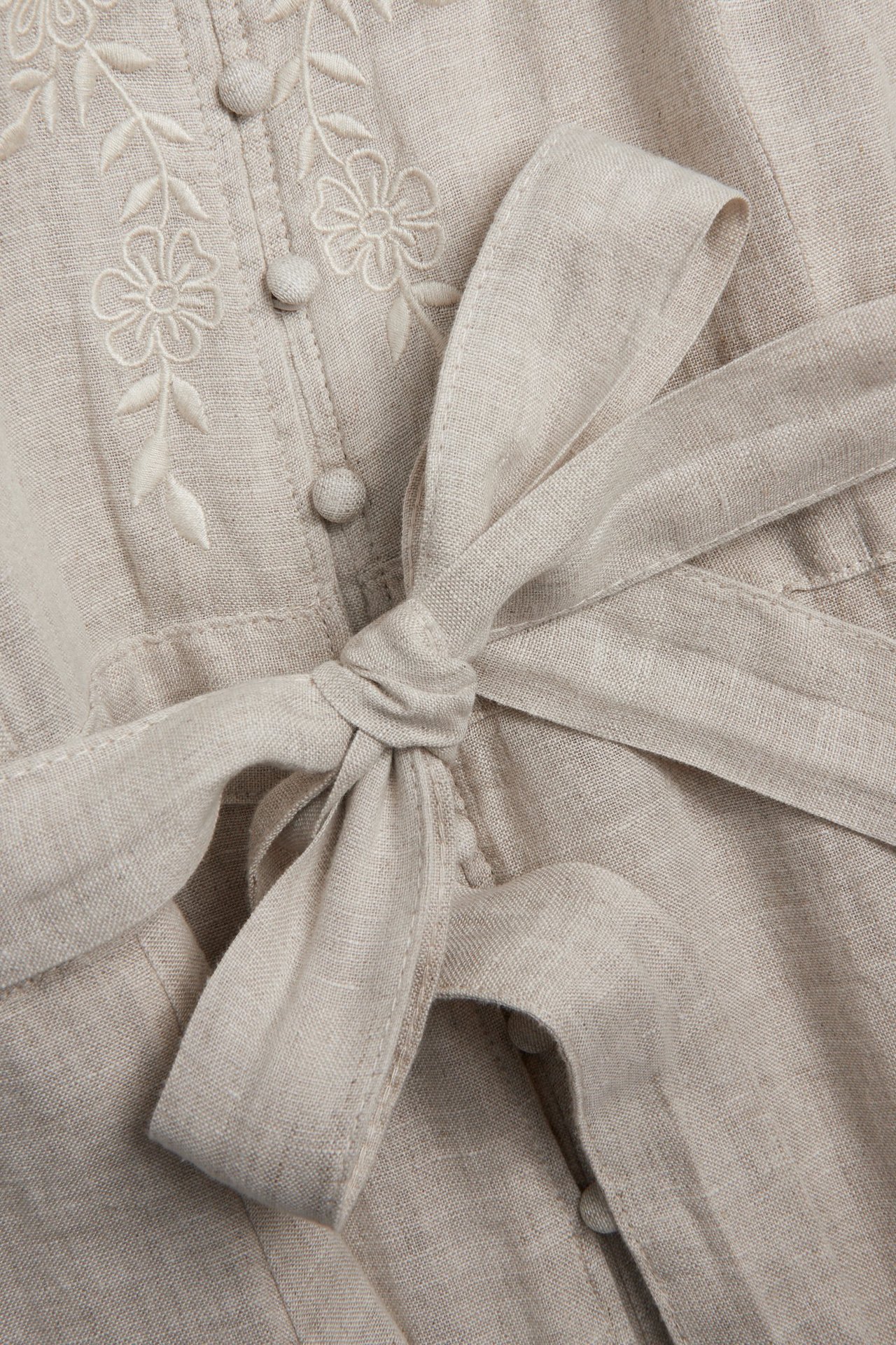 Close-up of a light beige linen dress with white floral embroidery, front buttons, and a matching tied belt.