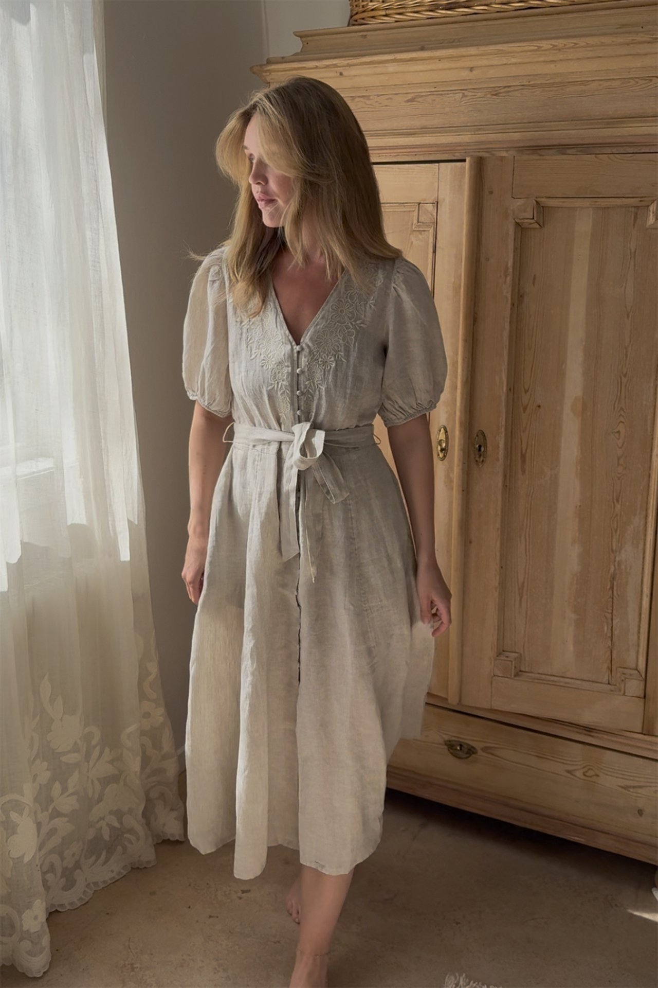 Vorderansicht einer Frau in einem hellbeigen Leinenkleid mit Stickereien, Puffärmeln und gebundenem Gürtel.