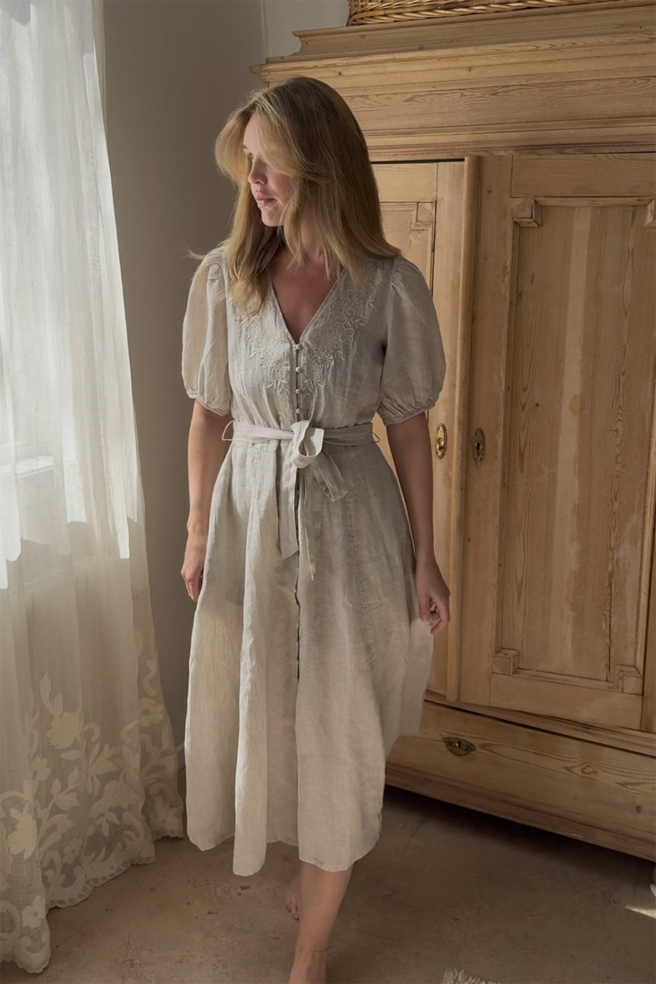 Front-side view of a woman in a light beige linen dress with embroidery, puff sleeves, and a tied belt.