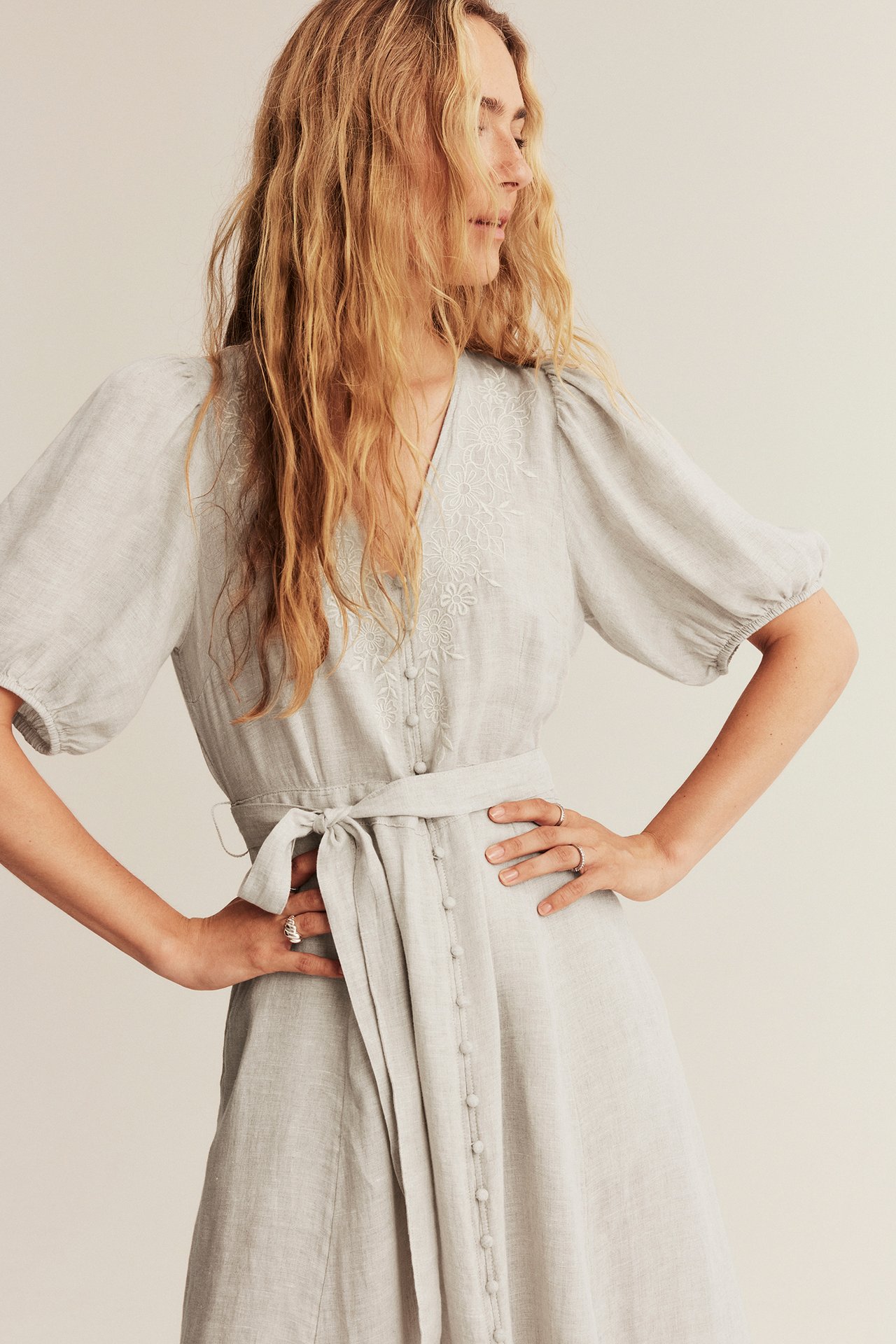 Woman wearing light gray linen dress with white floral embroidery, puff sleeves, self-tie belt, and rings.