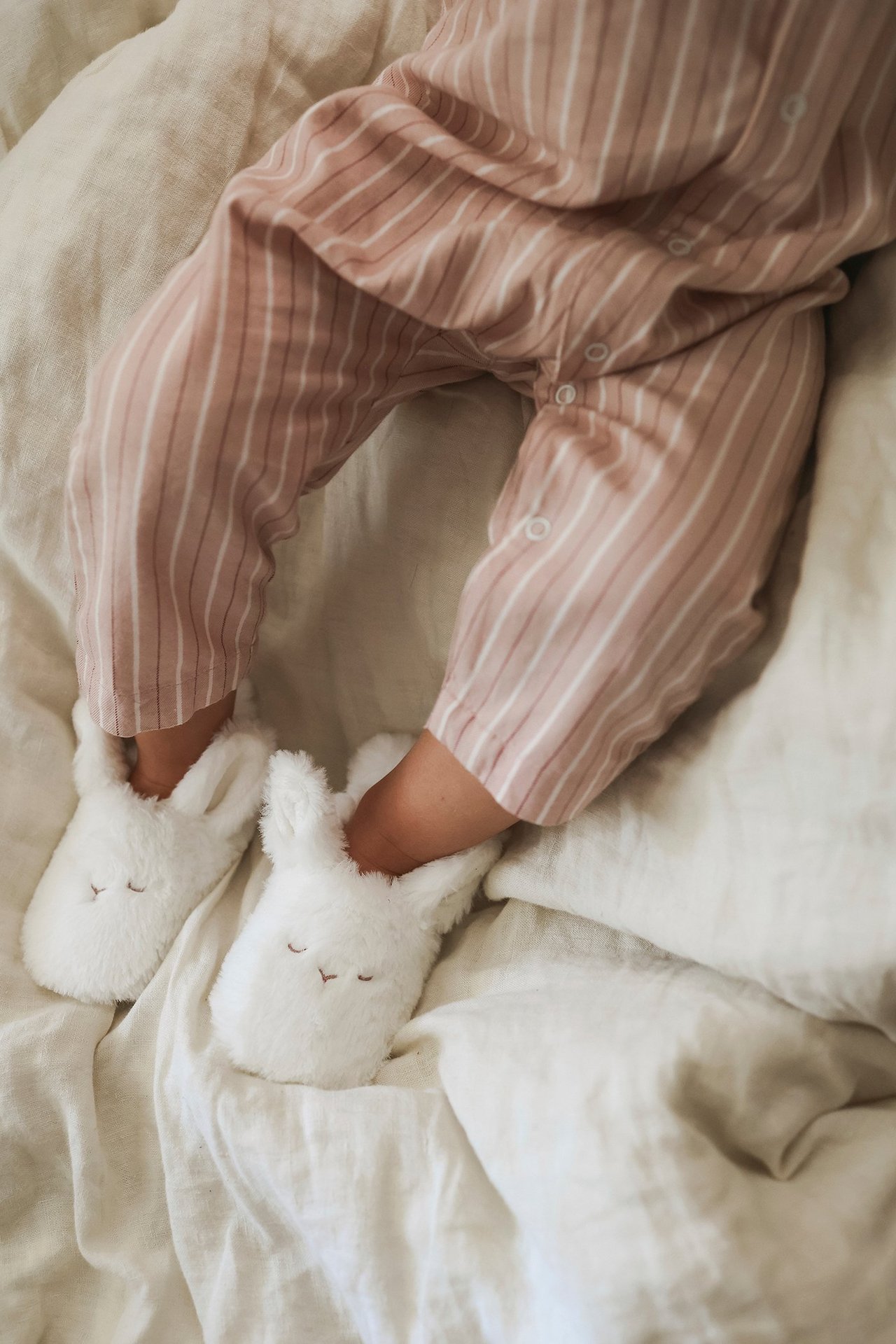 Draufsicht: Baby trägt cremeweiße Plüsch-Hasenhausschuhe und einen rosa-weiß gestreiften Strampler.