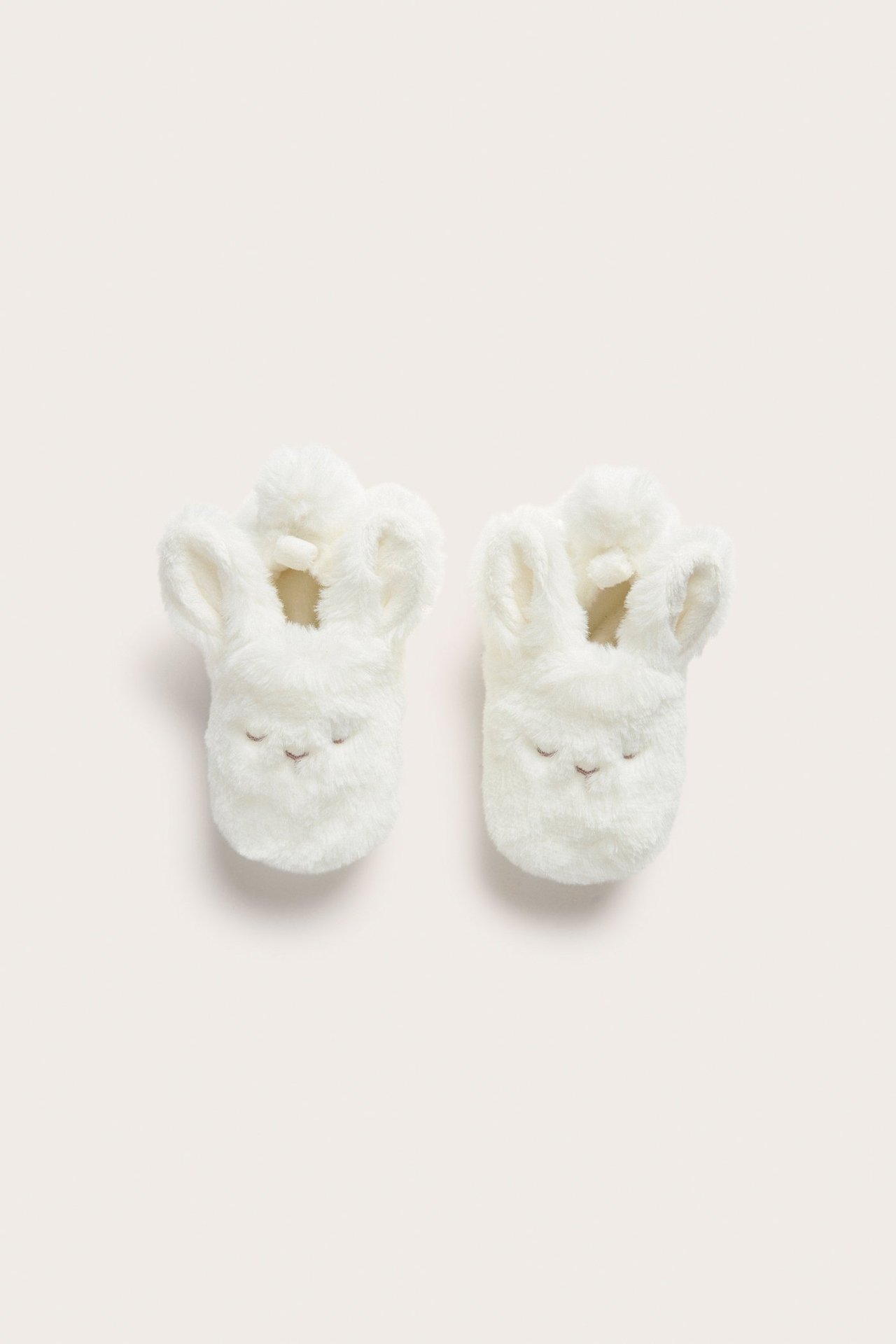 Top-down view of two offwhite pile baby slippers with rabbit ears and embroidered sleepy faces.