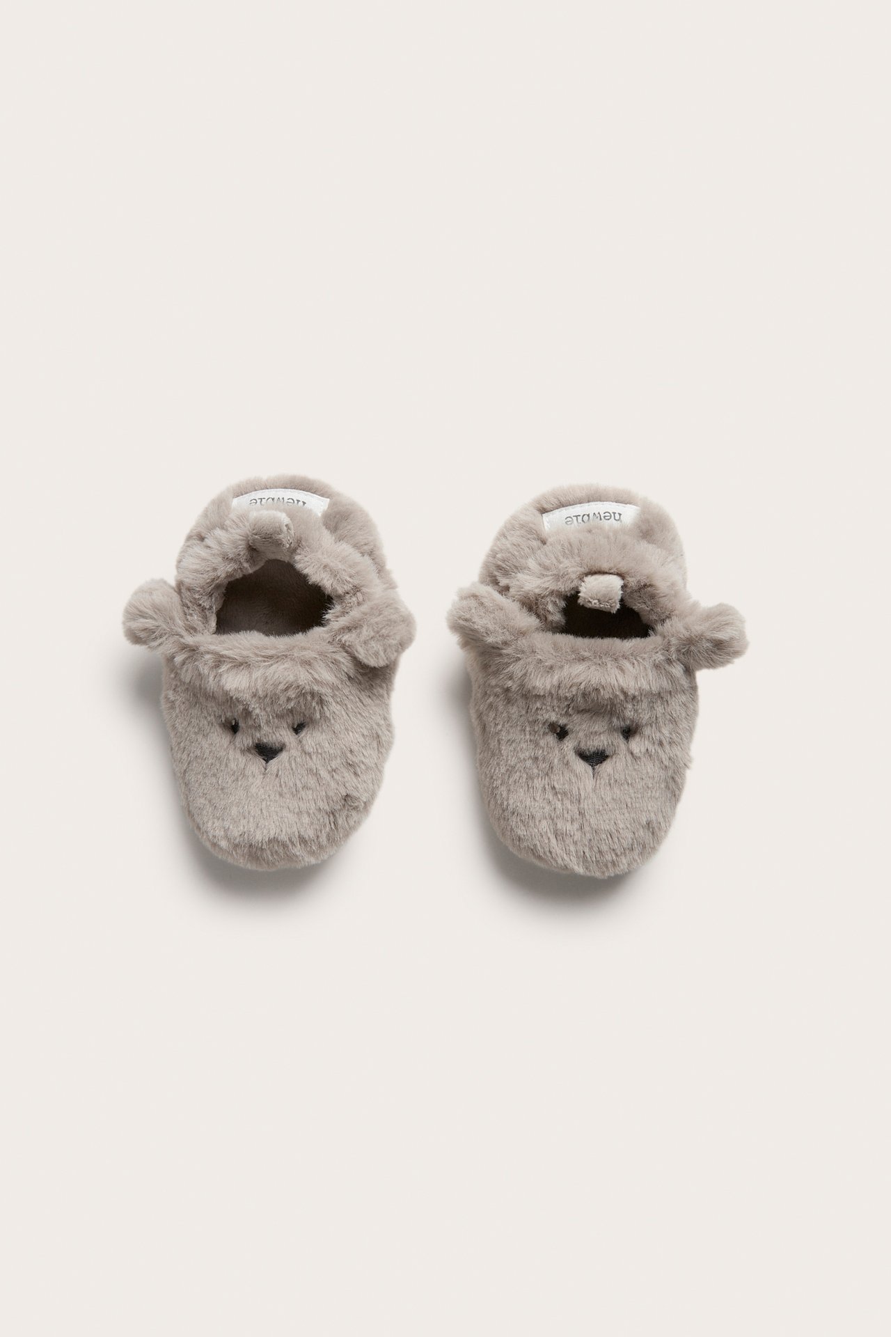 Draufsicht auf zwei beigefarbene flauschige Baby-Bärchenhausschuhe mit aufgestickten Gesichtern und Ohren.