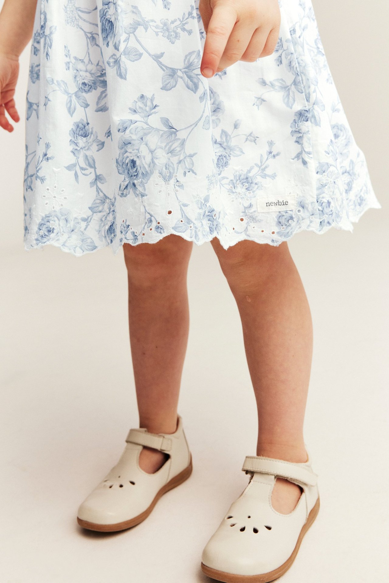 Kind in cremeweißem Blumenkleid mit blauen Blüten, besticktem Saum und beigen Mary Jane Schuhen. Vorderansicht.