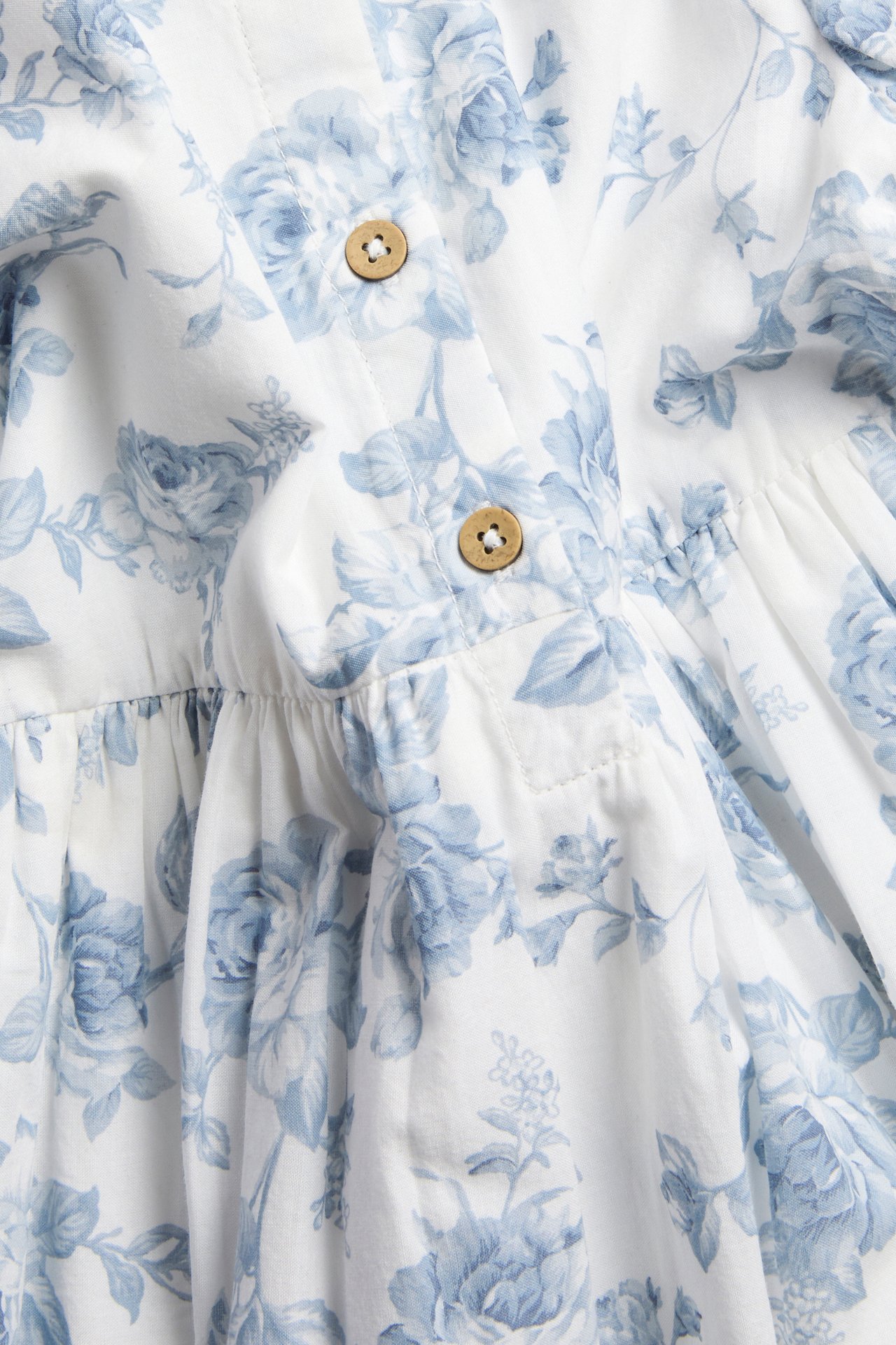 Nahaufnahme eines cremeweißen gewebten Kleides mit blauem Blumenmuster und hellbraunen Holzknöpfen.