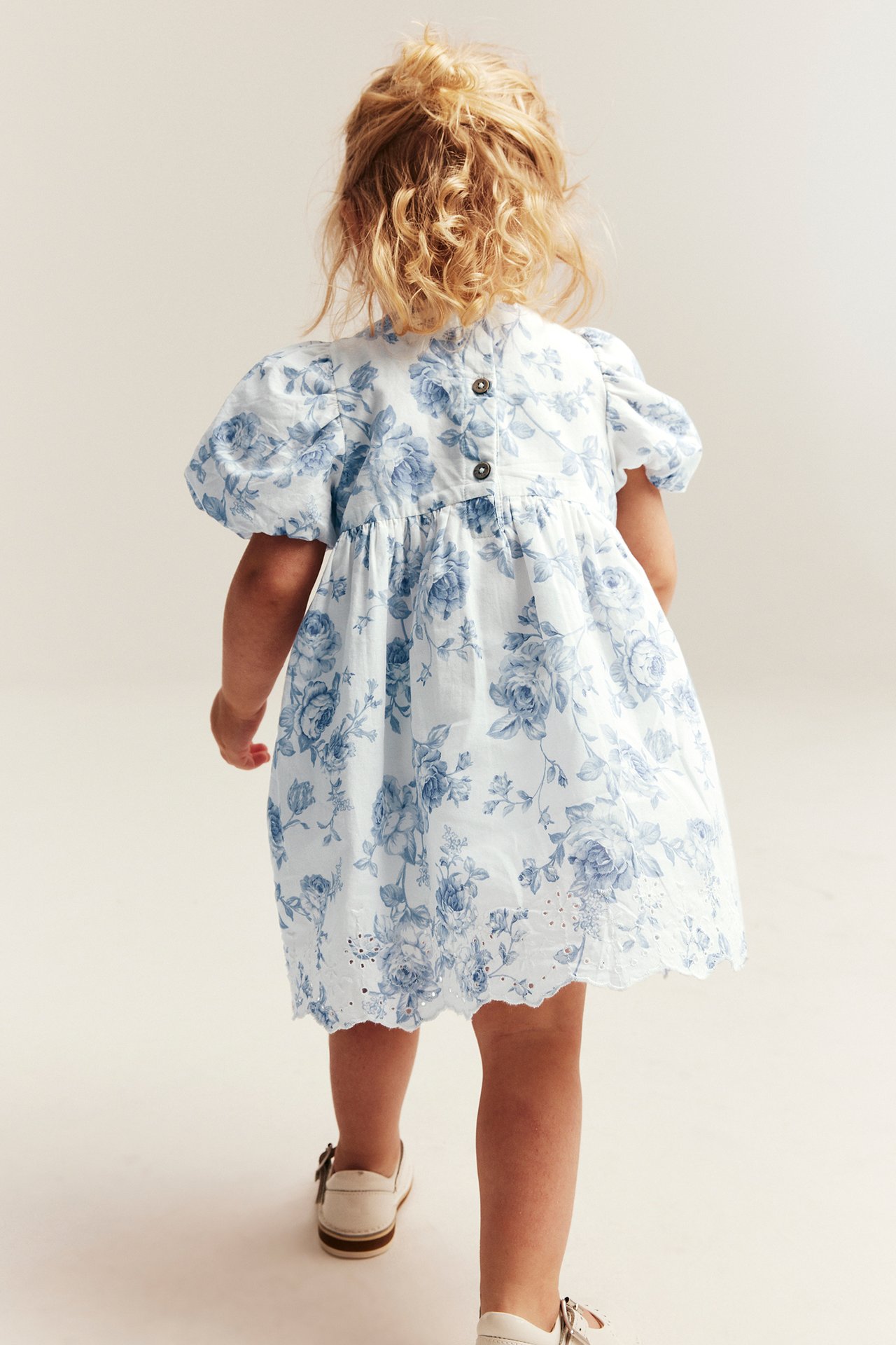 Back view: Kid's off-white floral dress with blue print, puff sleeves, back buttons, embroidered hem. White shoes.