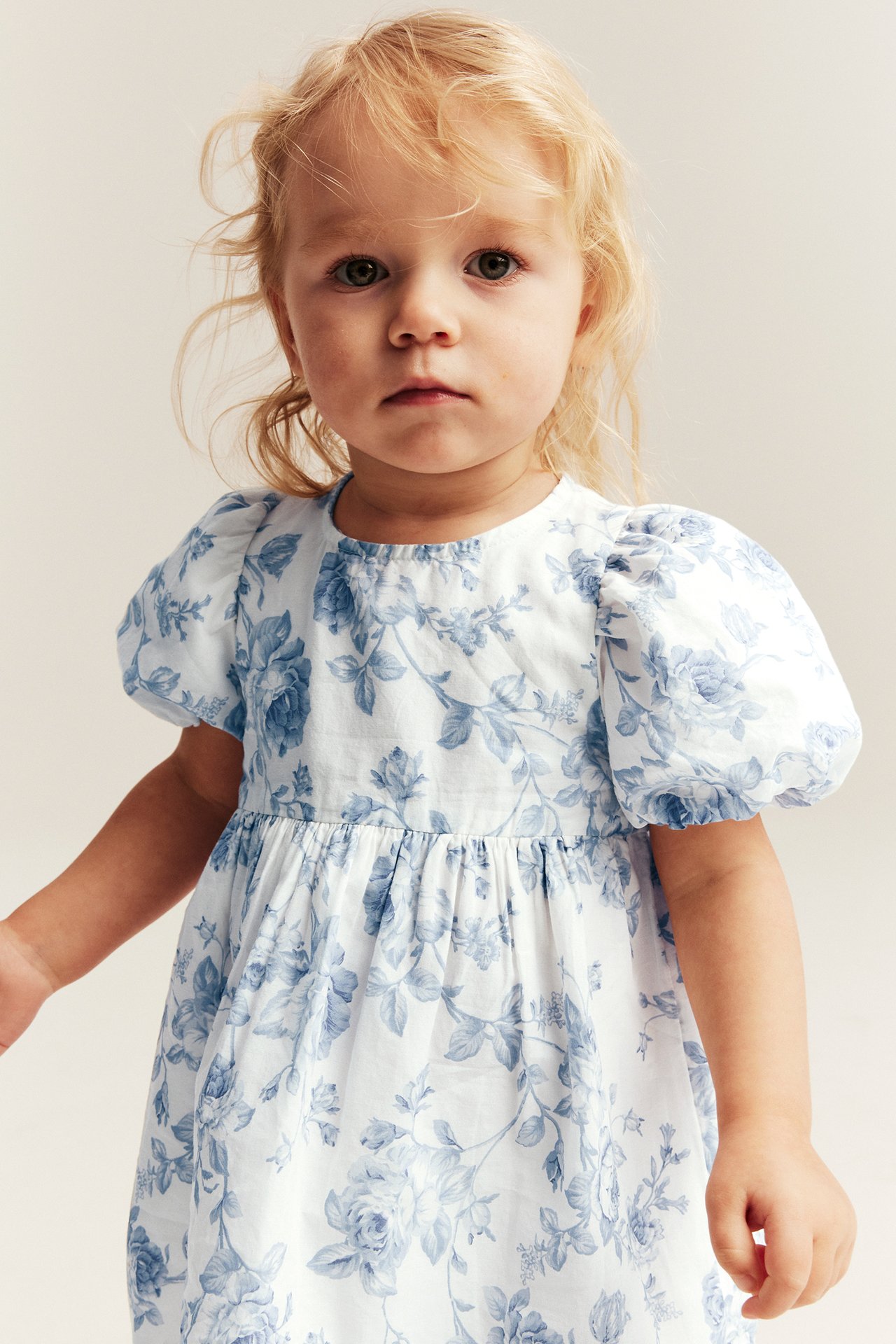 Front view of a kid wearing an off-white dress with blue floral print and puffed sleeves.