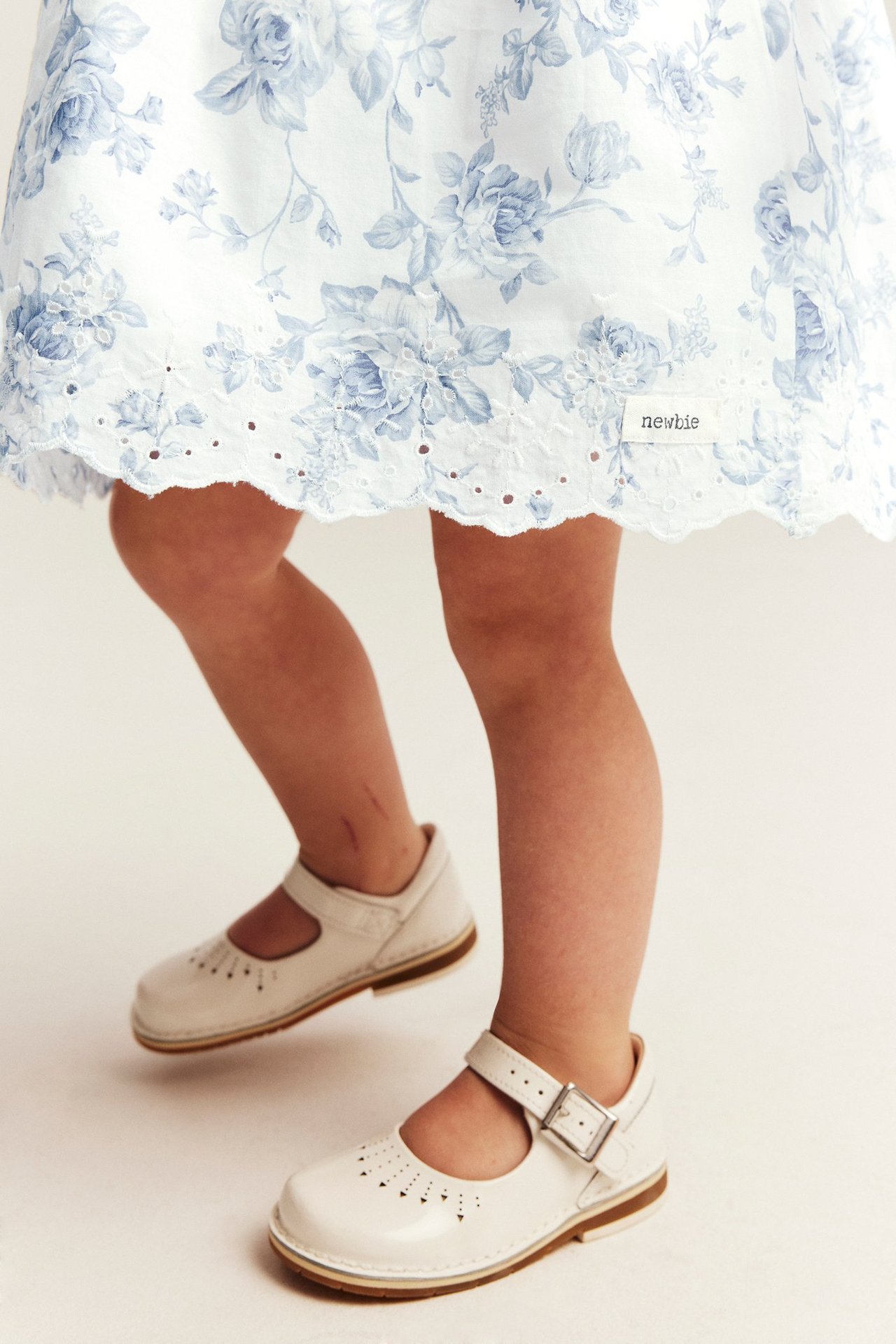 Vorderansicht von unten: Kind in weißem Blumenkleid mit Stickerei-Saum und weißen Mary Jane Schuhen.
