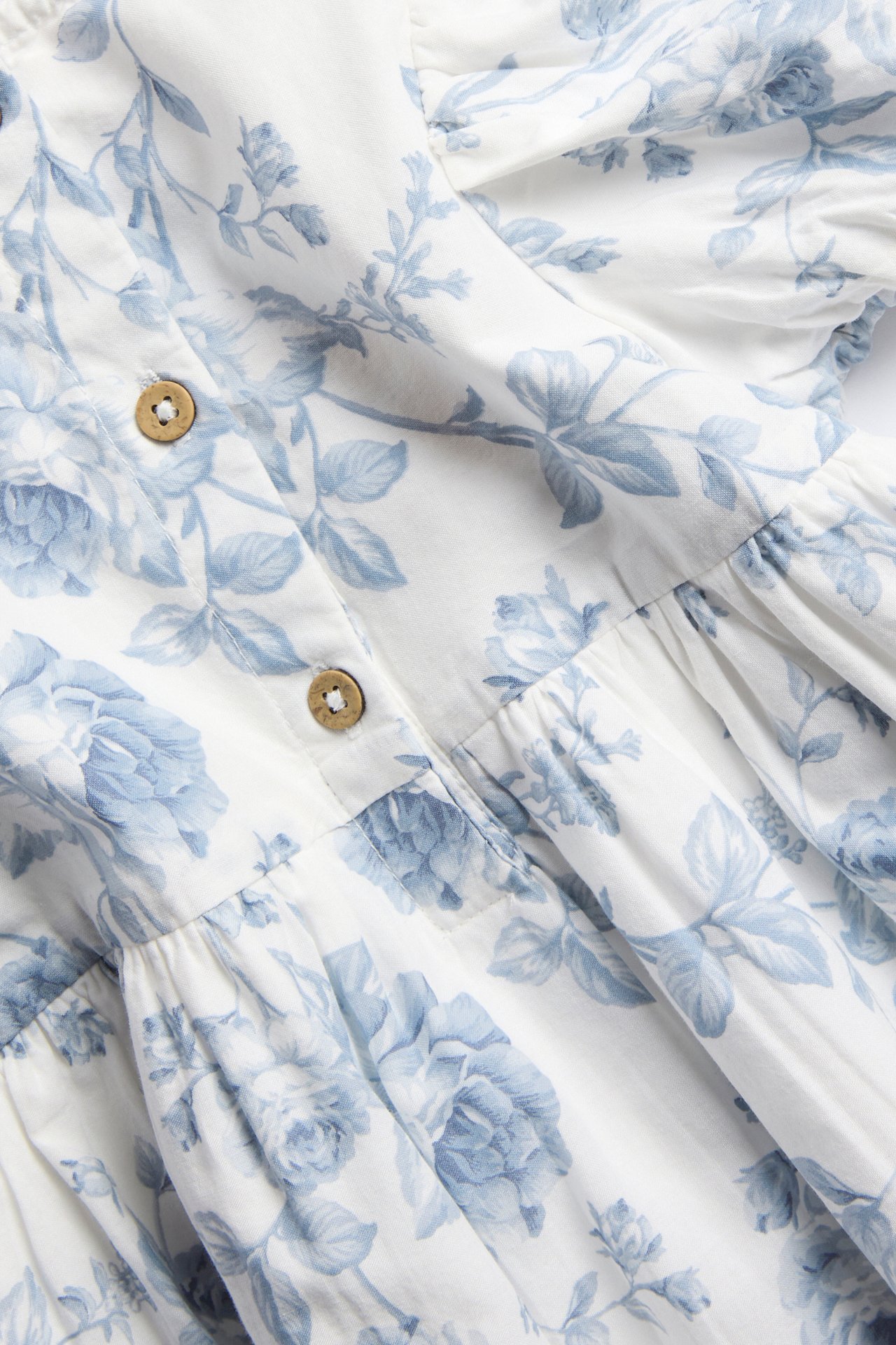 Nahaufnahme eines cremeweißen Damenkleides mit hellblauem Blumenmuster, das zwei Holzknöpfe und gerafften Stoff zeigt.