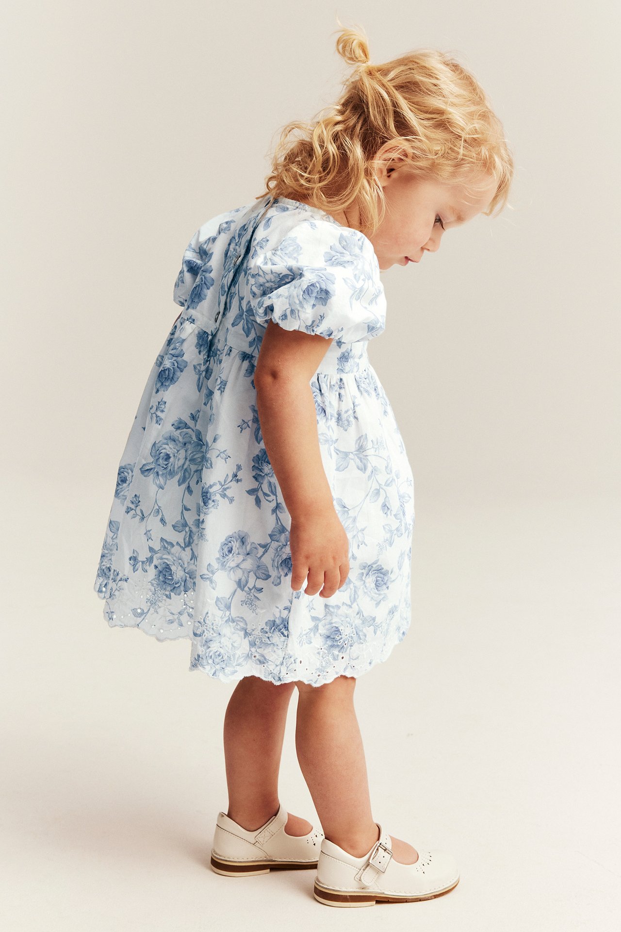 Side view of a kid wearing an off-white floral dress with puff sleeves, an embroidered hem, and off-white Mary Jane shoes.