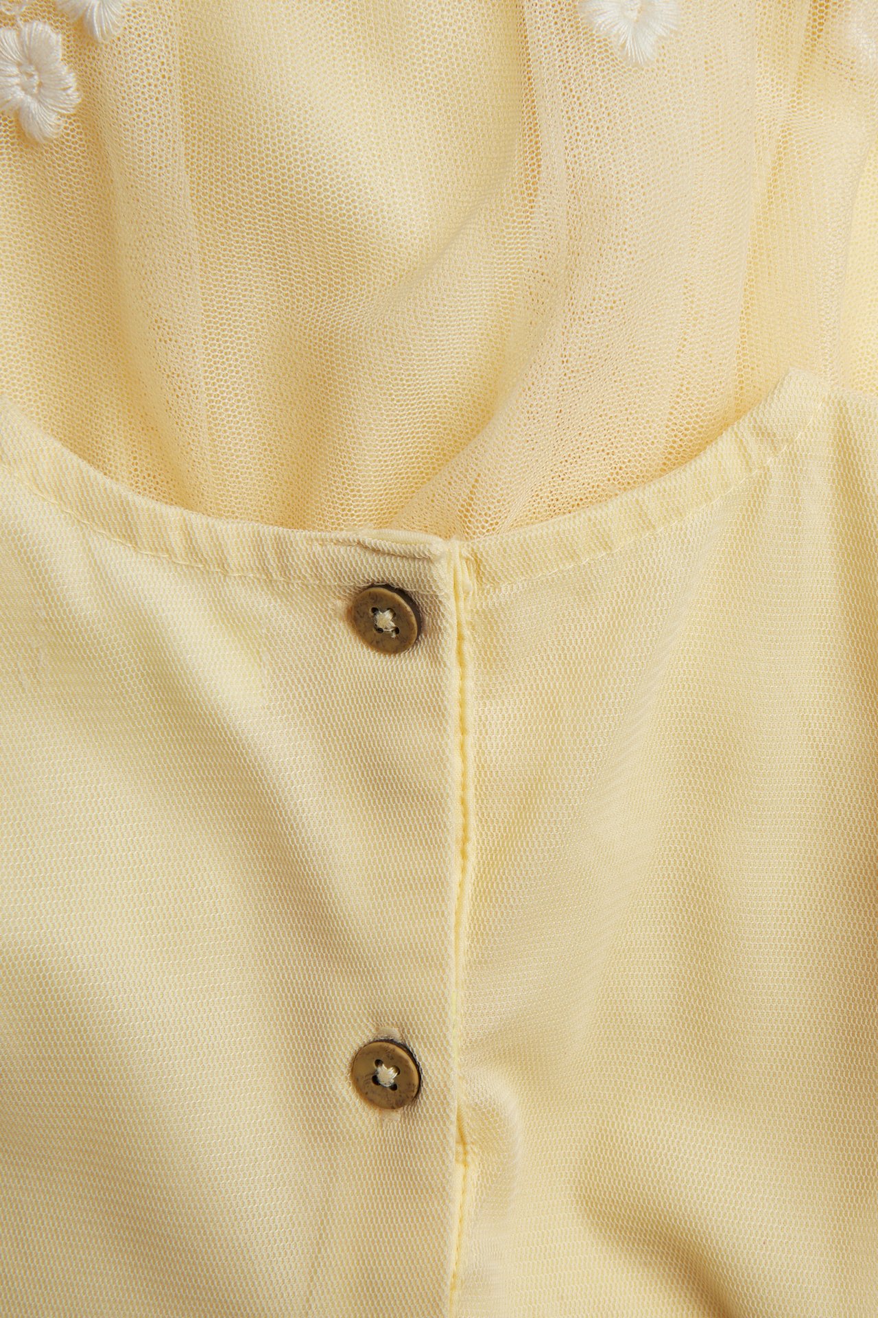 Nahaufnahme eines gelben Mesh-Kleides mit weißer Blumenstickerei und zwei braunen Knöpfen.