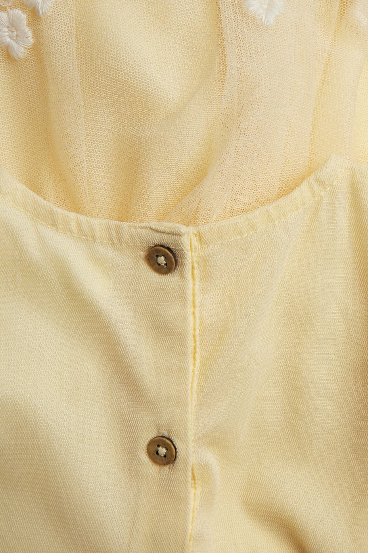 Nahaufnahme eines gelben Mesh-Kleides mit weißer Blumenstickerei und zwei braunen Knöpfen.