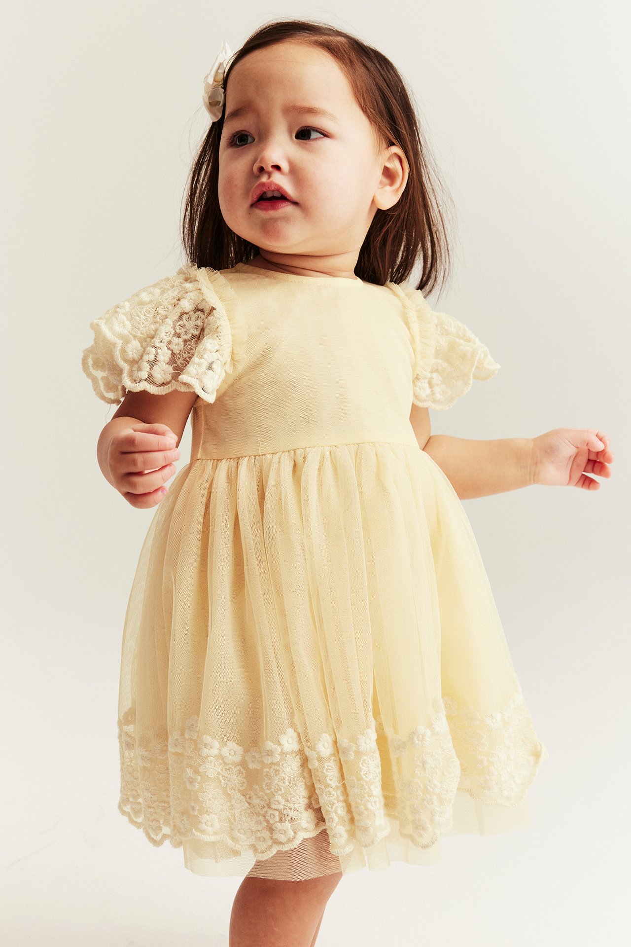 Front view of a baby girl in a yellow mesh dress with lace embroidery and a white hair clip.