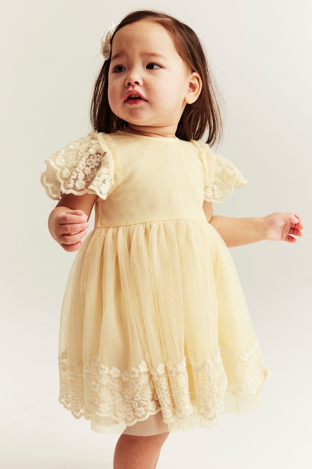 Front view of a baby girl in a yellow mesh dress with lace embroidery and a white hair clip.