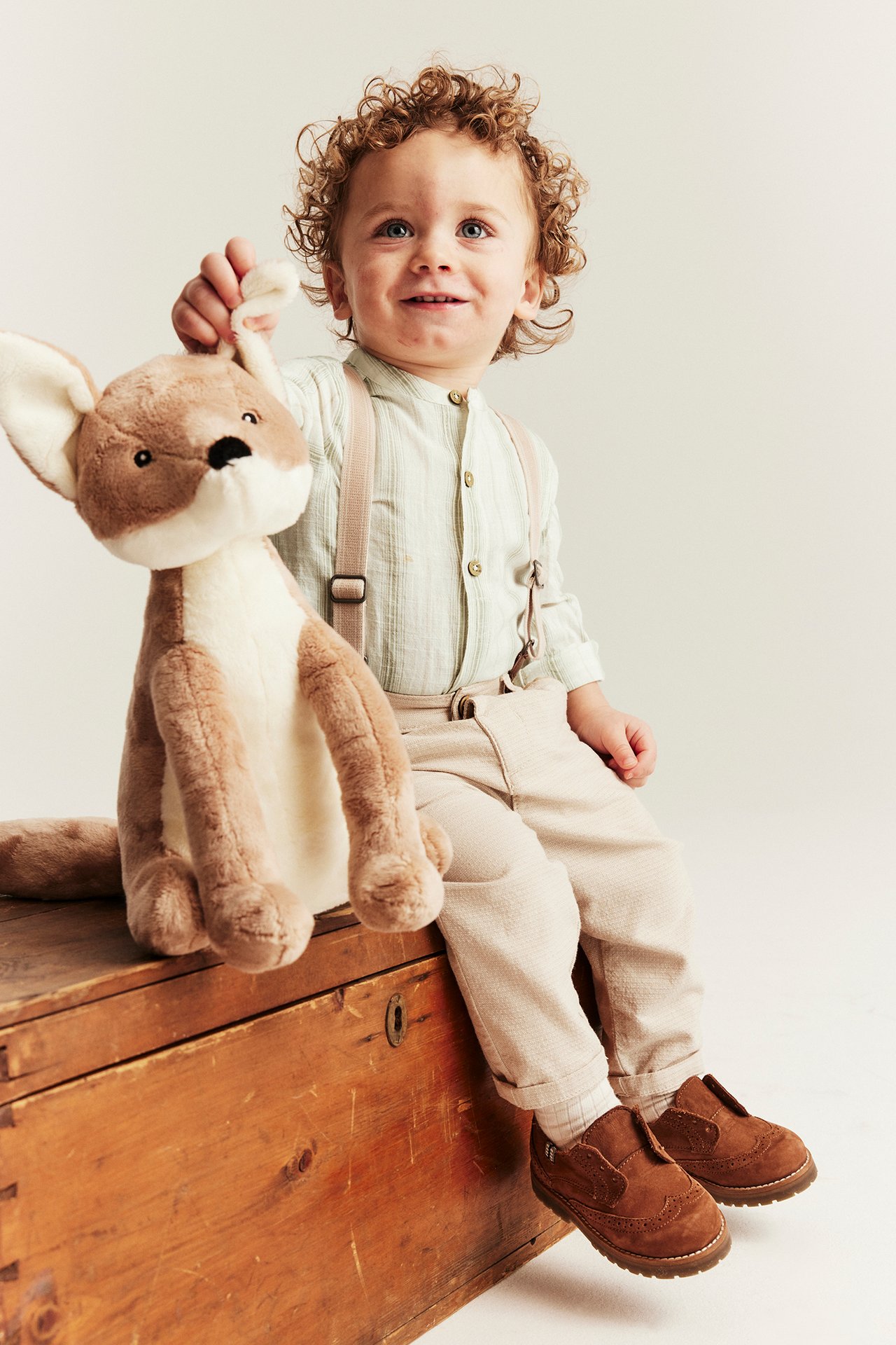 Vorderansicht eines Babys in einem hellgrün gestreiften Langarm-Shirtbody, beigen Hosen, Hosenträgern, braunen Schuhen, hält ein Känguru-Spielzeug.