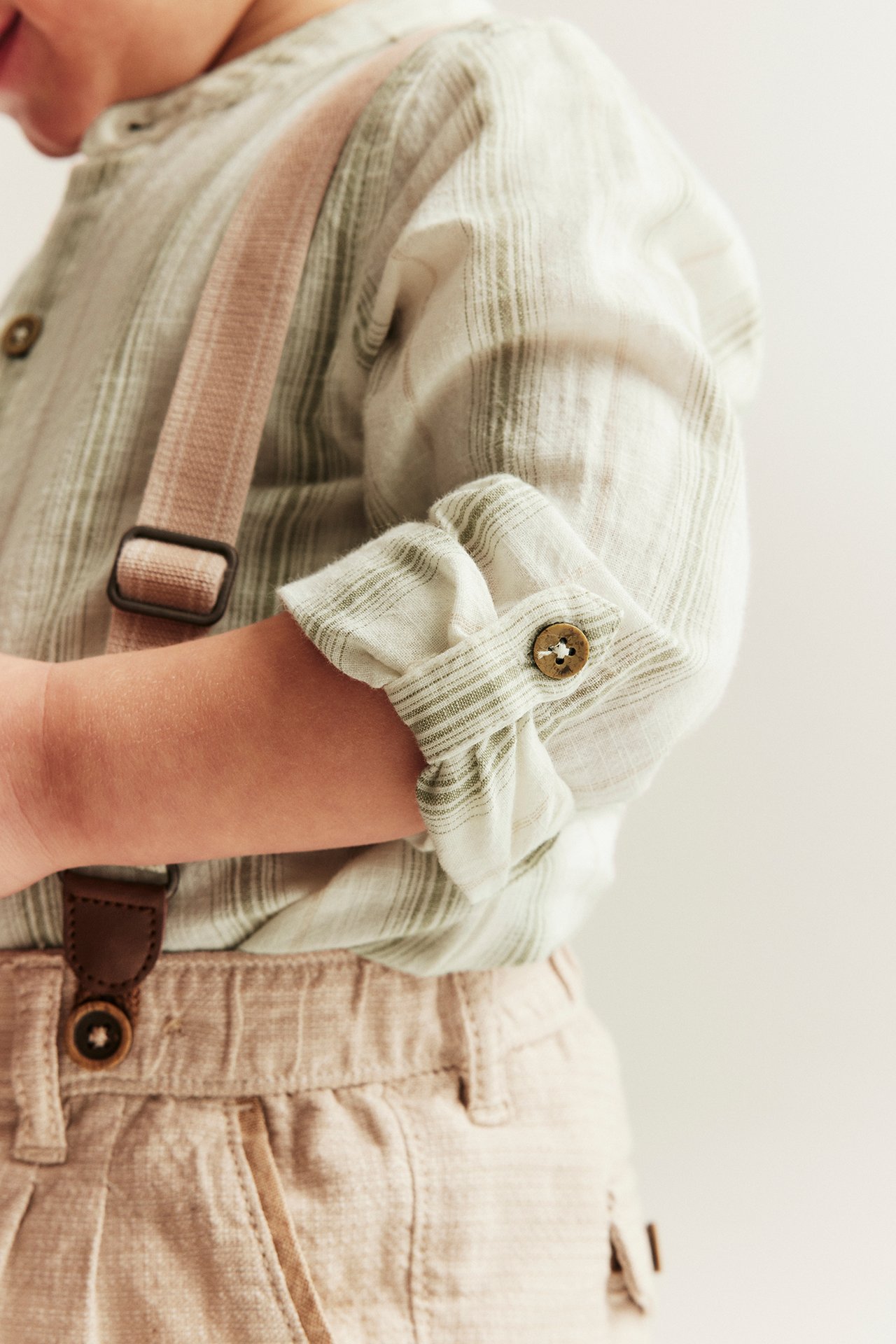 Close-up of a baby wearing a light green striped shirt body with rolled-up sleeves, beige pants, and pink suspenders.