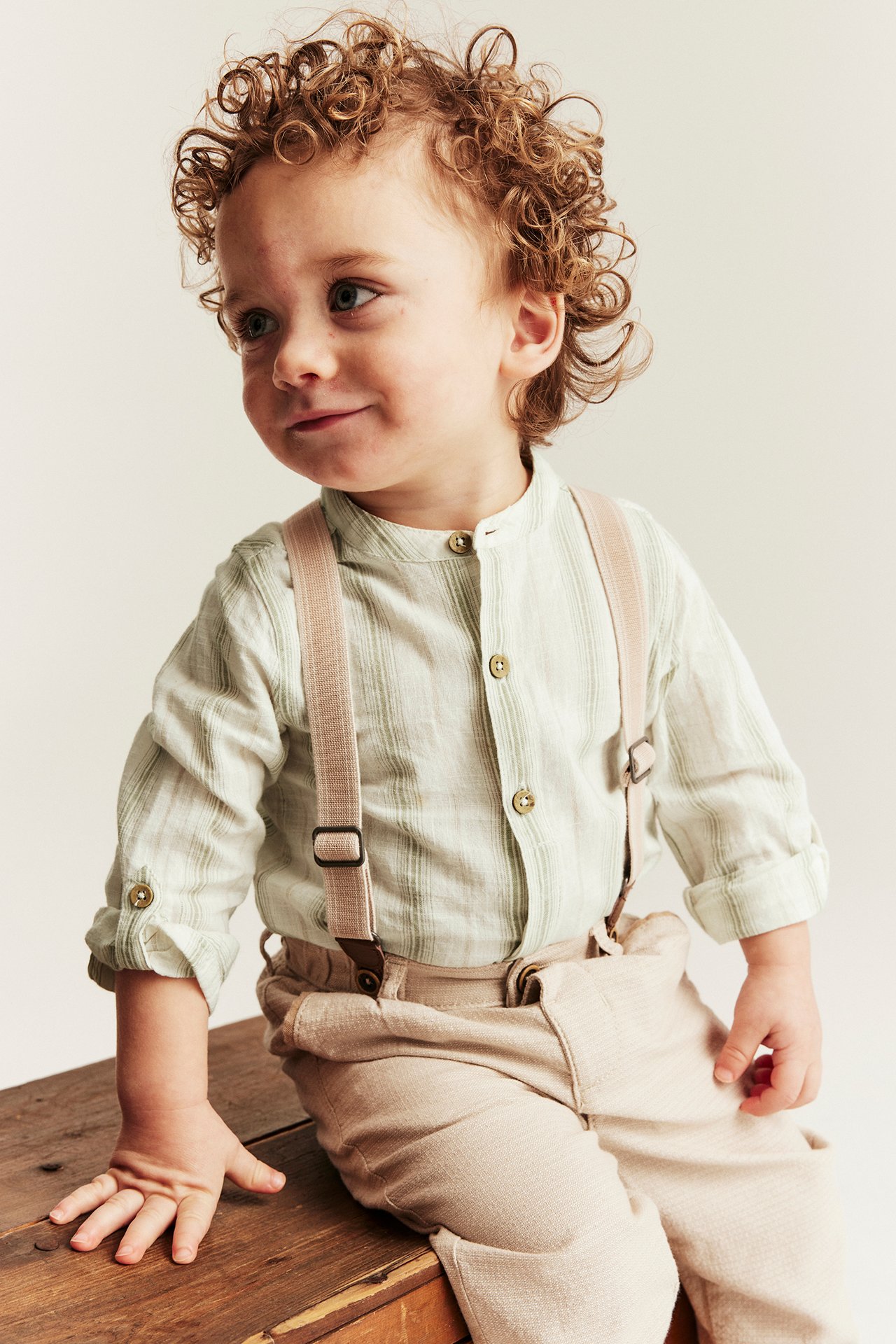 Vorderansicht: Baby in hellgrün gestreiftem Langarm-Shirtbody, beiger Hose und Hosenträgern.