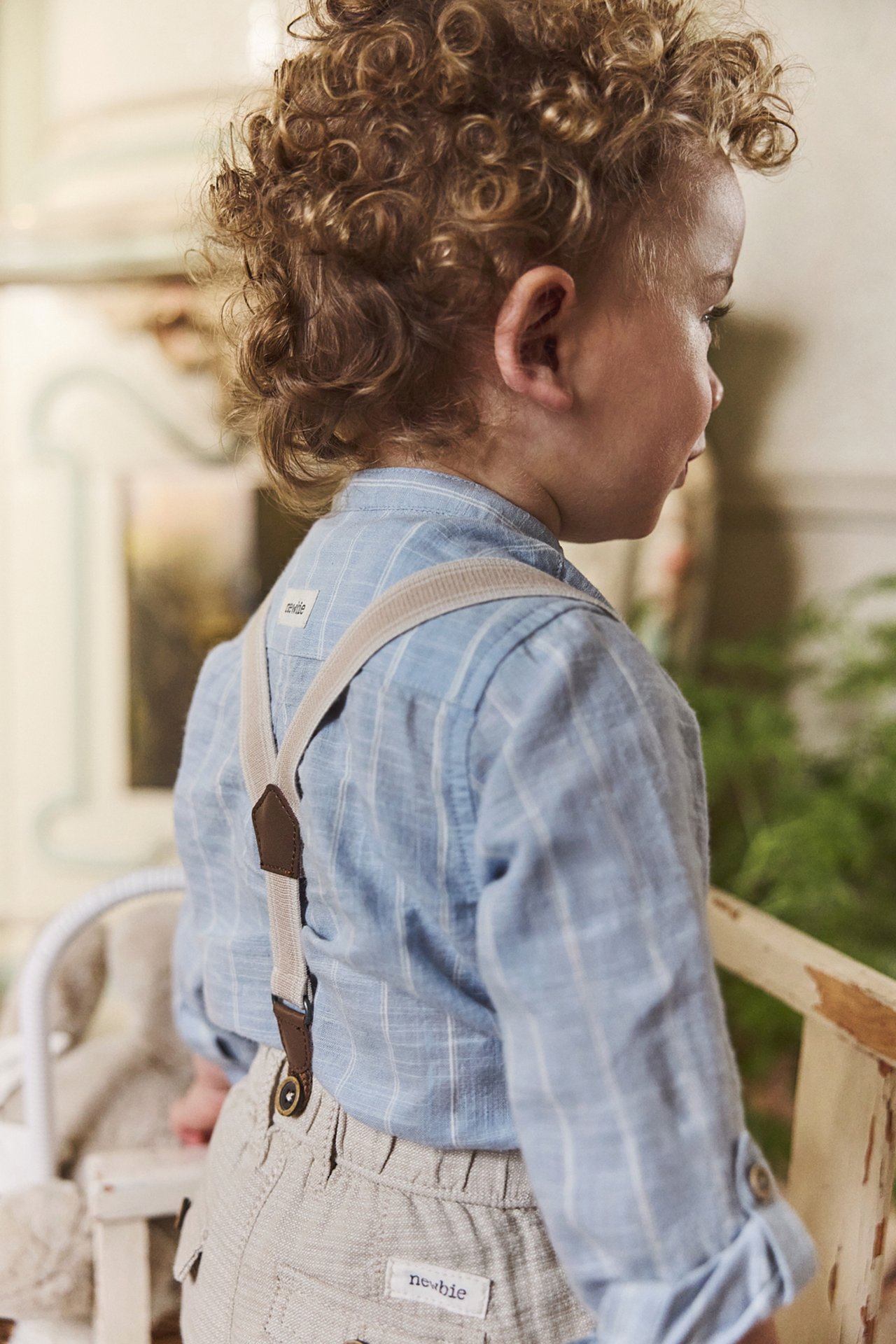 Back view: Kid in blue striped long-sleeved shirt body, beige trousers, and suspenders.