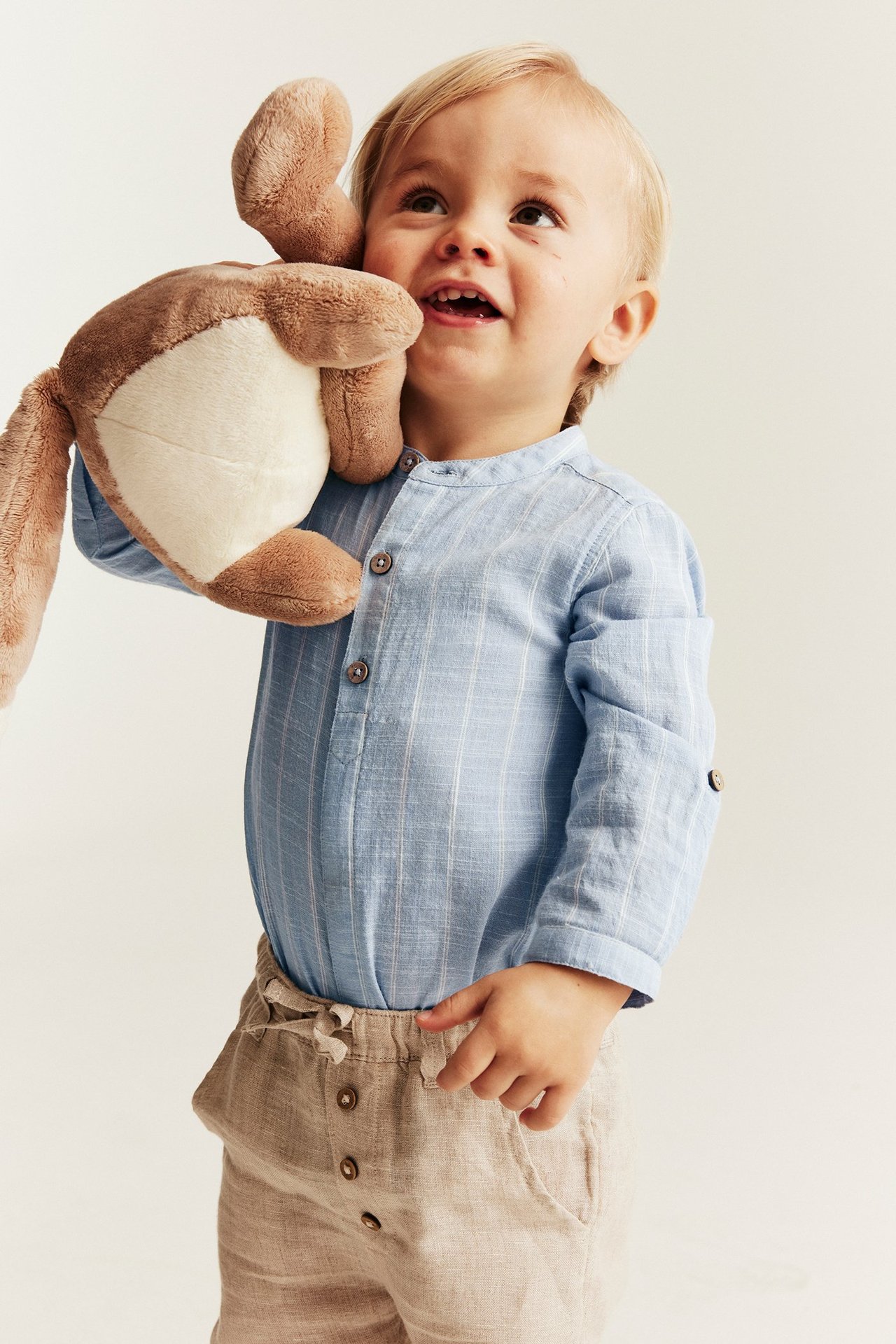 Babyjunge trägt einen blau gestreiften Langarm-Shirt-Body und beigefarbene Shorts, hält ein braunes Plüschtier.