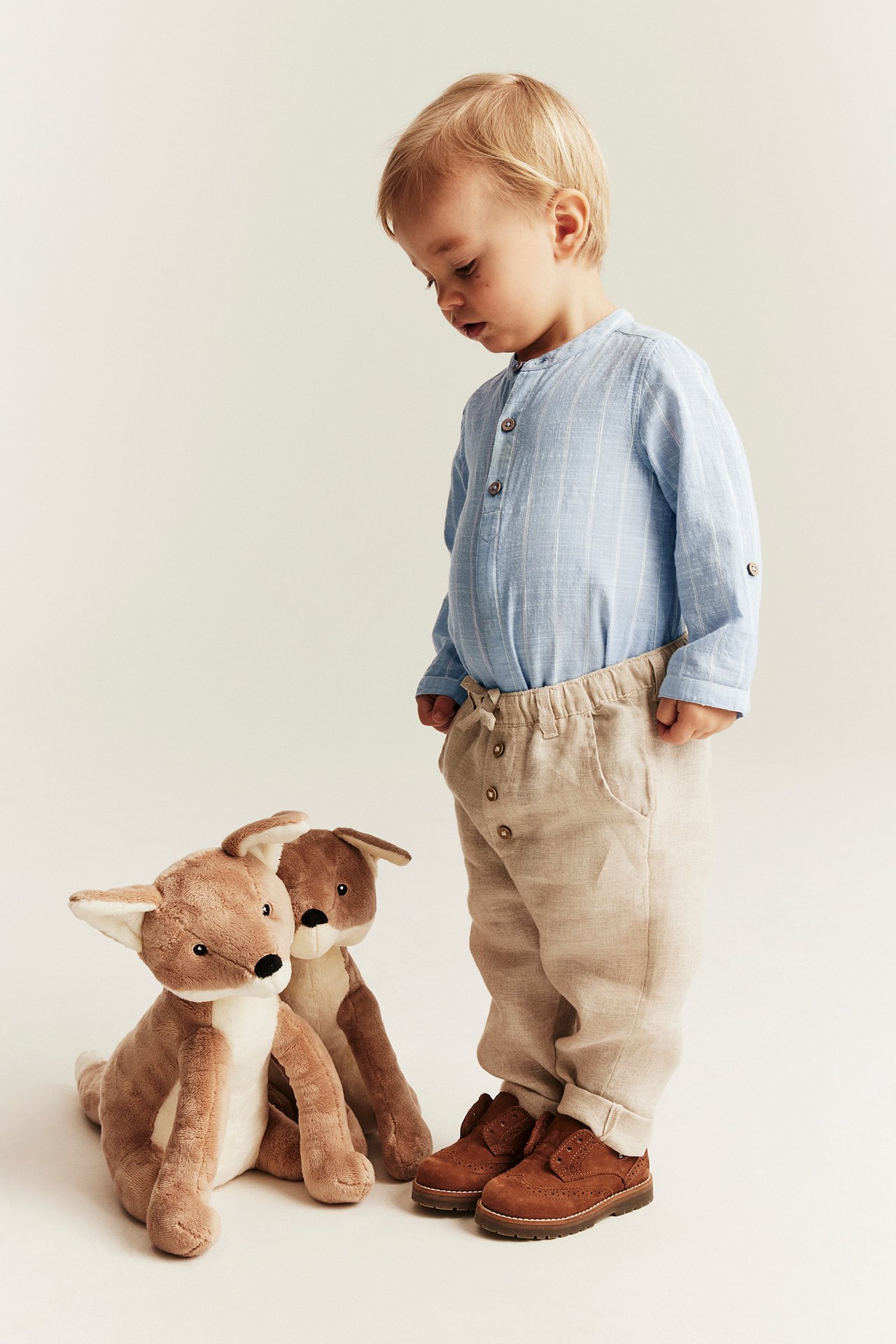 Front view of a baby wearing a light blue striped long-sleeved shirt bodysuit, beige trousers, and brown shoes.