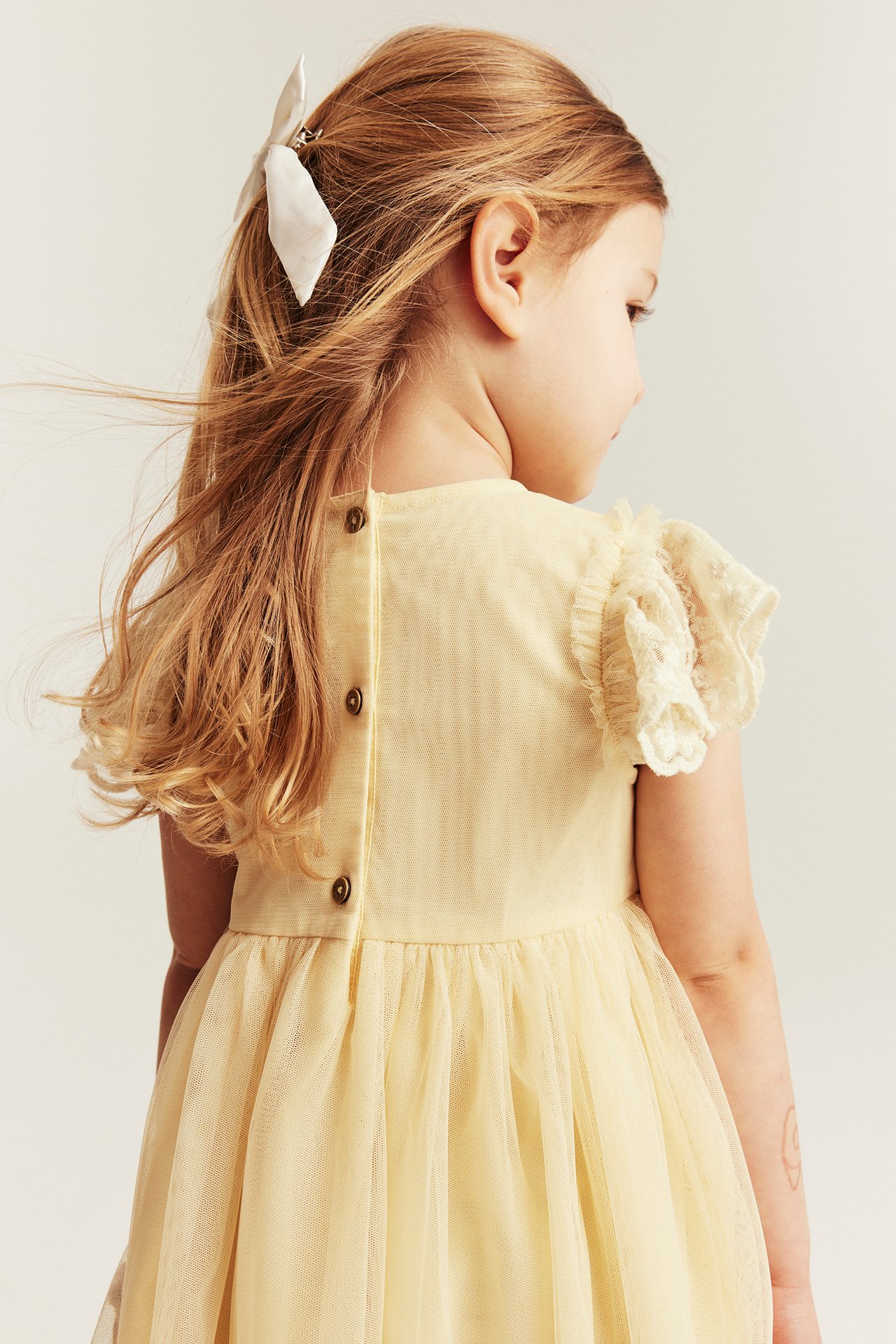Back view: Kid in light yellow mesh dress with back buttons, ruffled sleeves, and a white bow clip.
