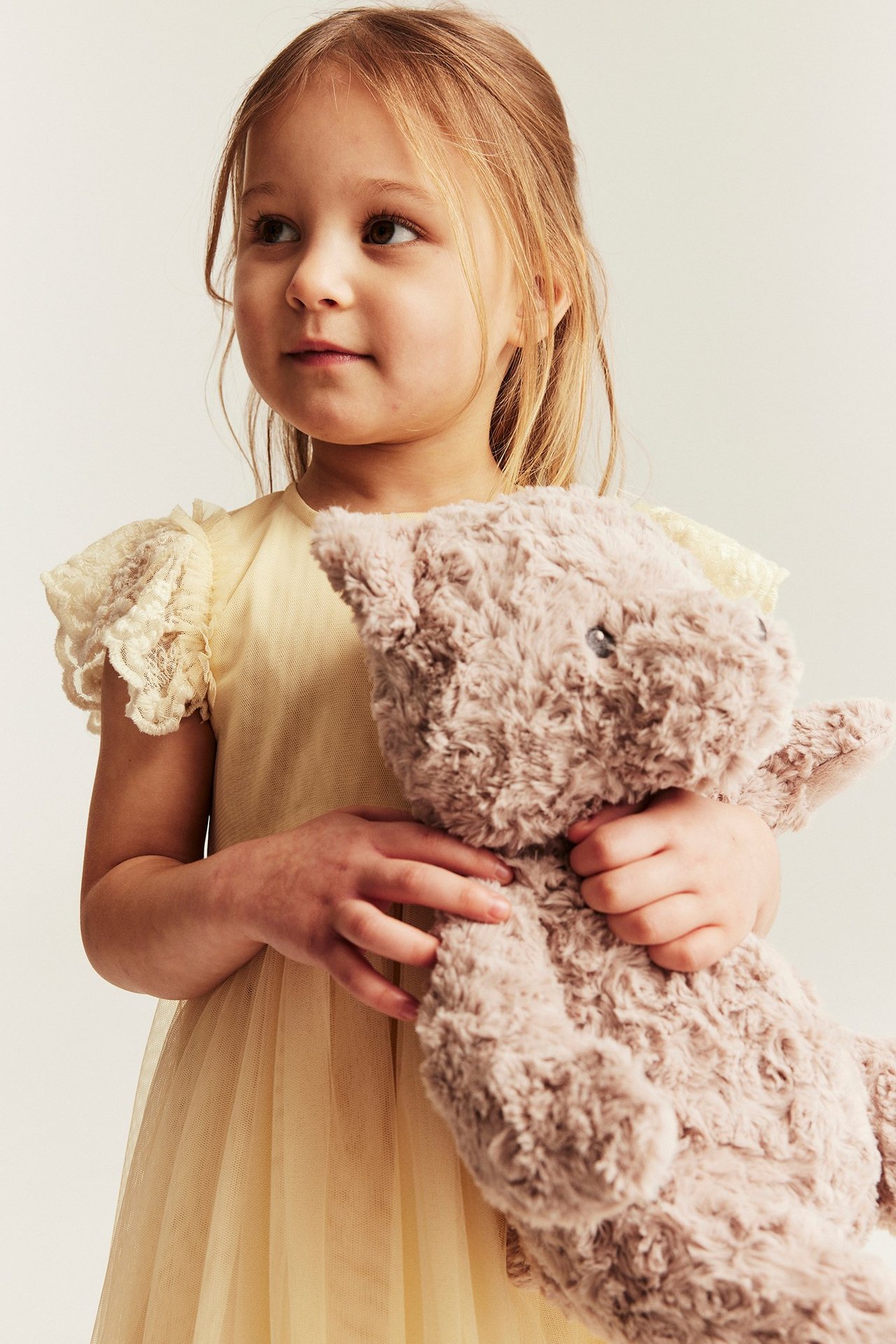 Front view of a kid in a yellow mesh dress with embroidered sleeves, holding a brown teddy bear.