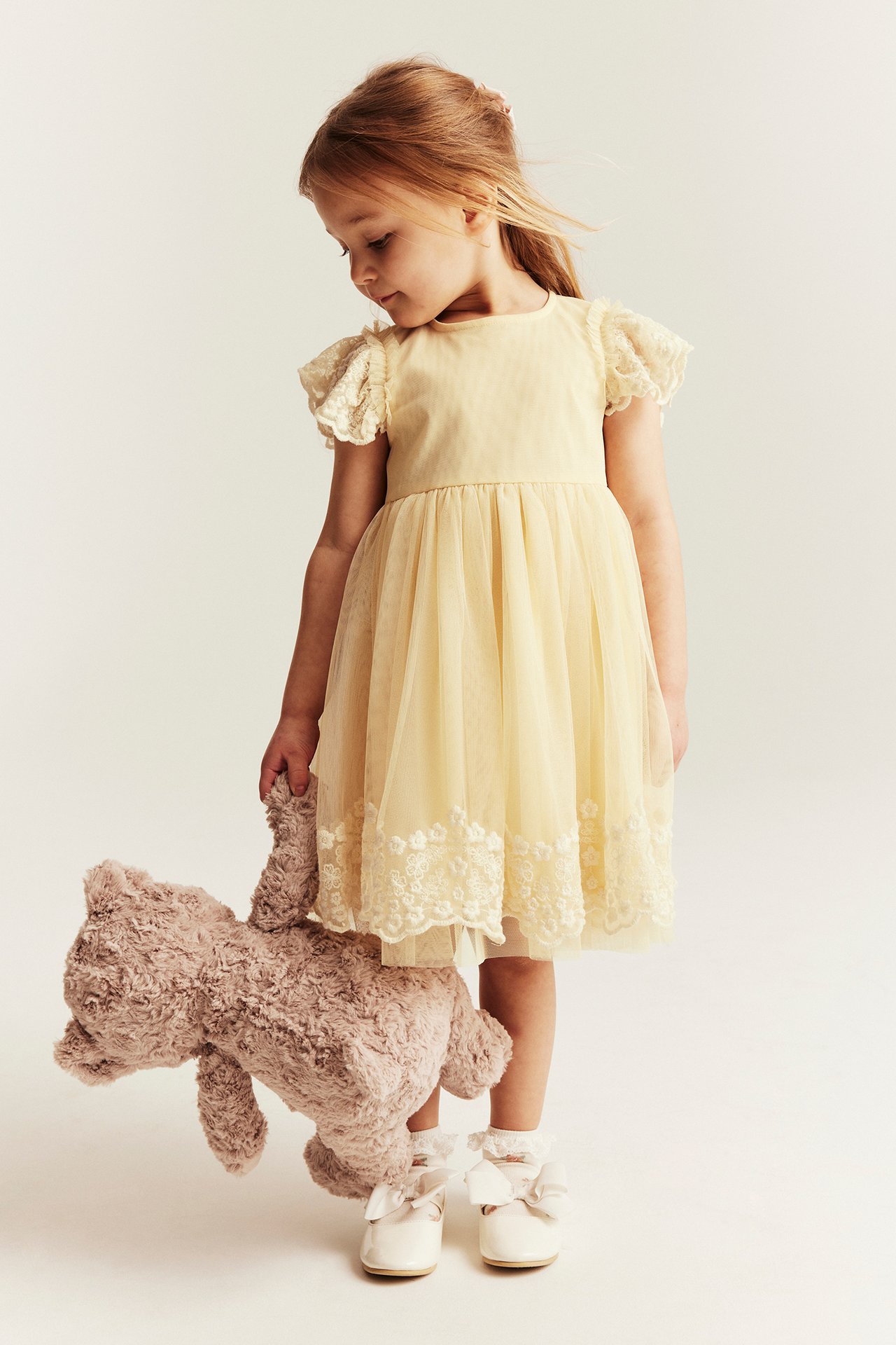 Front view of a kid wearing a yellow mesh embroidered dress, white bow shoes, and holding a brown teddy bear.
