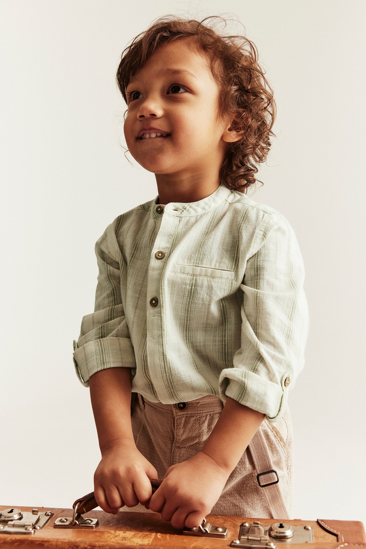 Vorderansicht: Kind in hellgrün gestreiftem Baumwollhemd mit Stehkragen, hochgekrempelten Ärmeln, beiger Hose, hält Koffer.