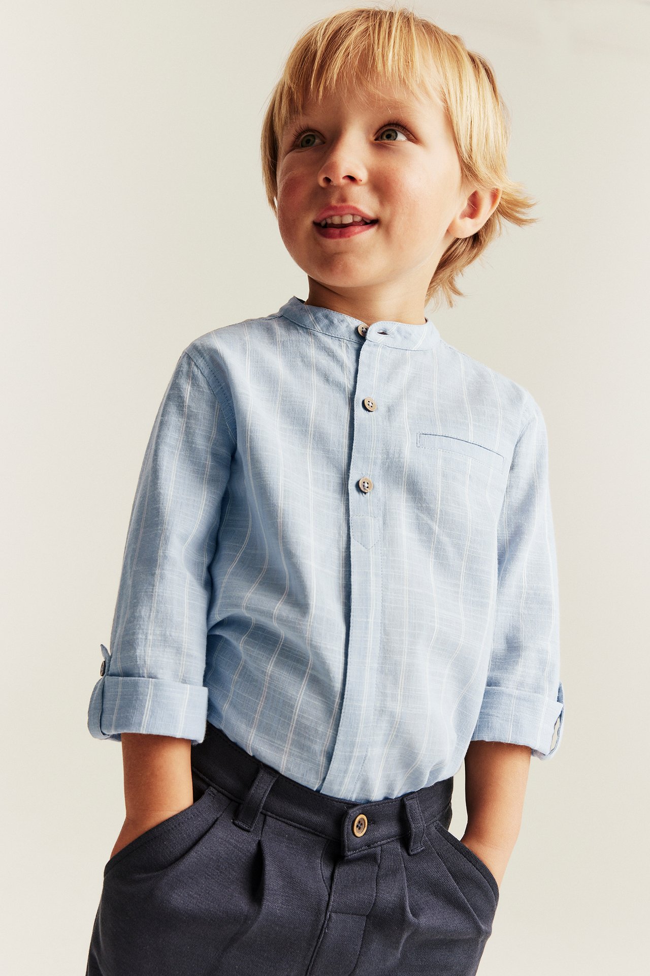 Front view: Kid in light blue striped band-collar shirt with rolled sleeves and dark blue trousers.