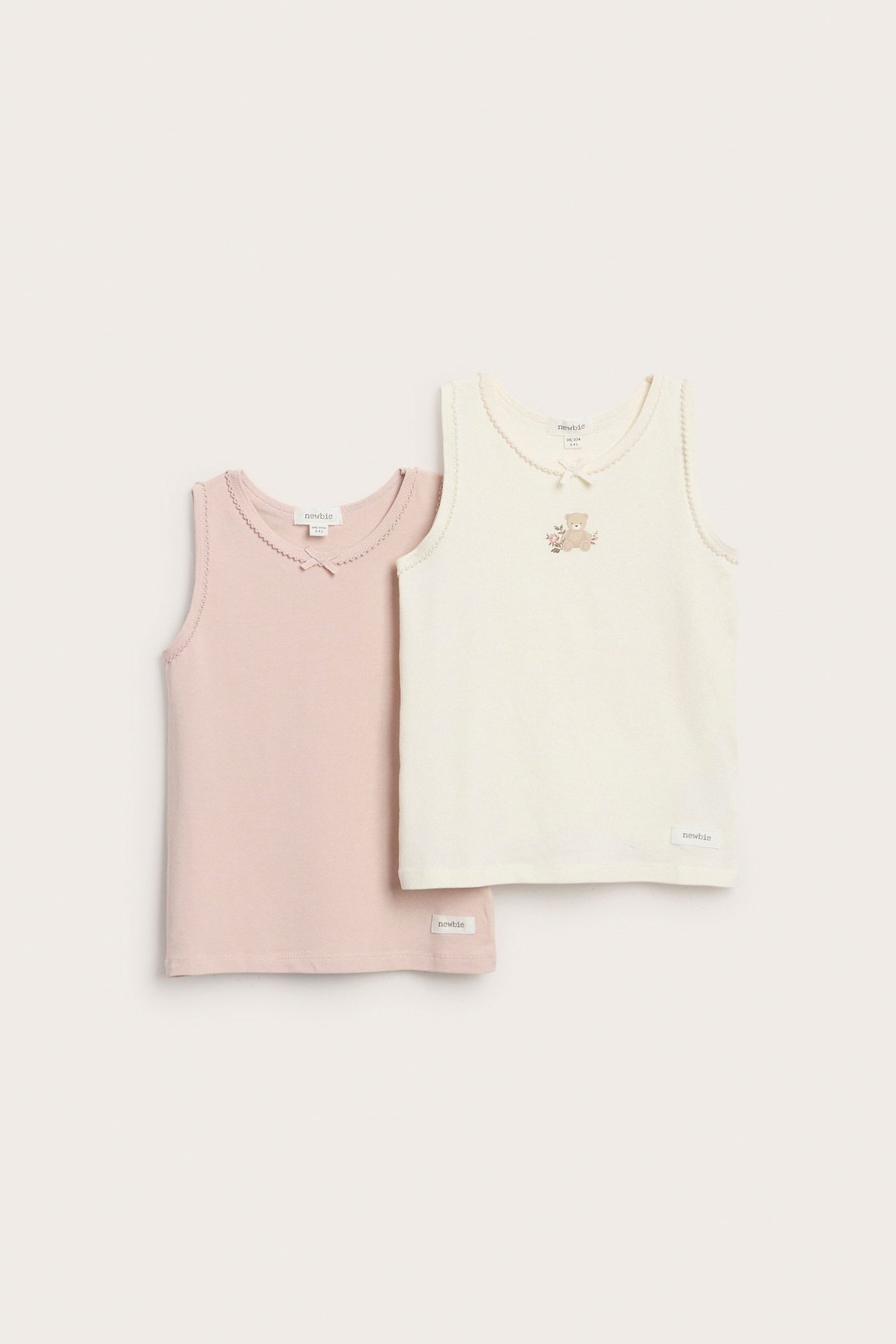 Zwei einfarbige Kinder-Tanktops, creme und rosa, mit Schleifen und Spitze.