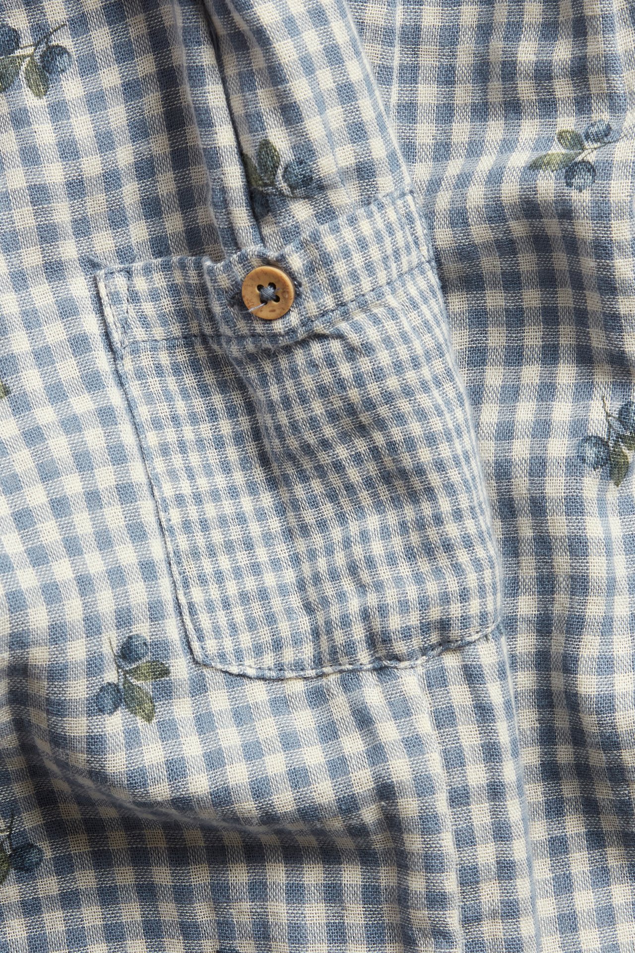 Nahaufnahme: Blau-weiß karierter Baumwollstoff mit Heidelbeer-Print, einer aufgesetzten Tasche und Holzknopf.