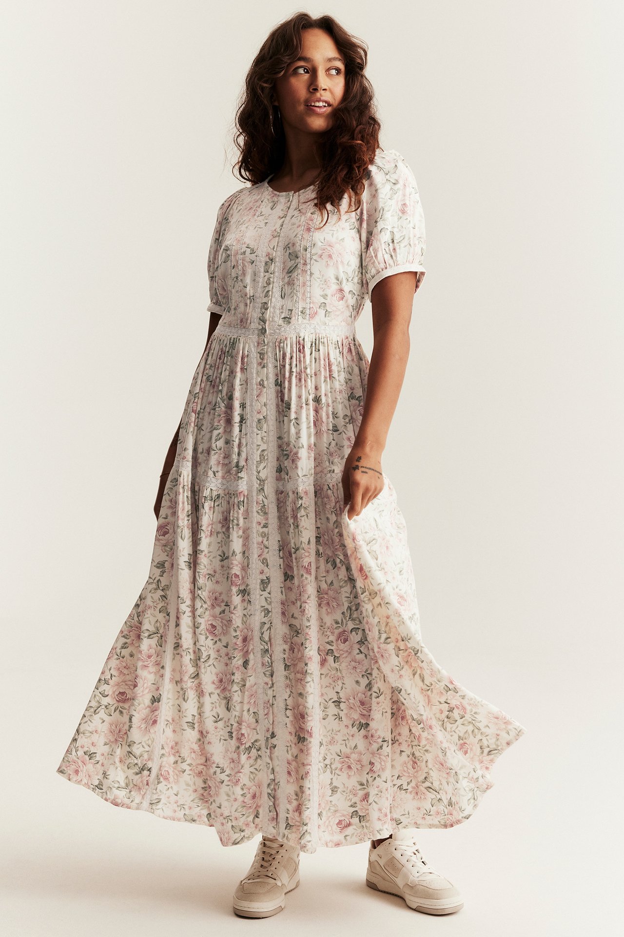 Vorderansicht: Frau in cremefarbenem Blumen-Maxikleid mit Puffärmeln, Spitze und beigen Sneakern.