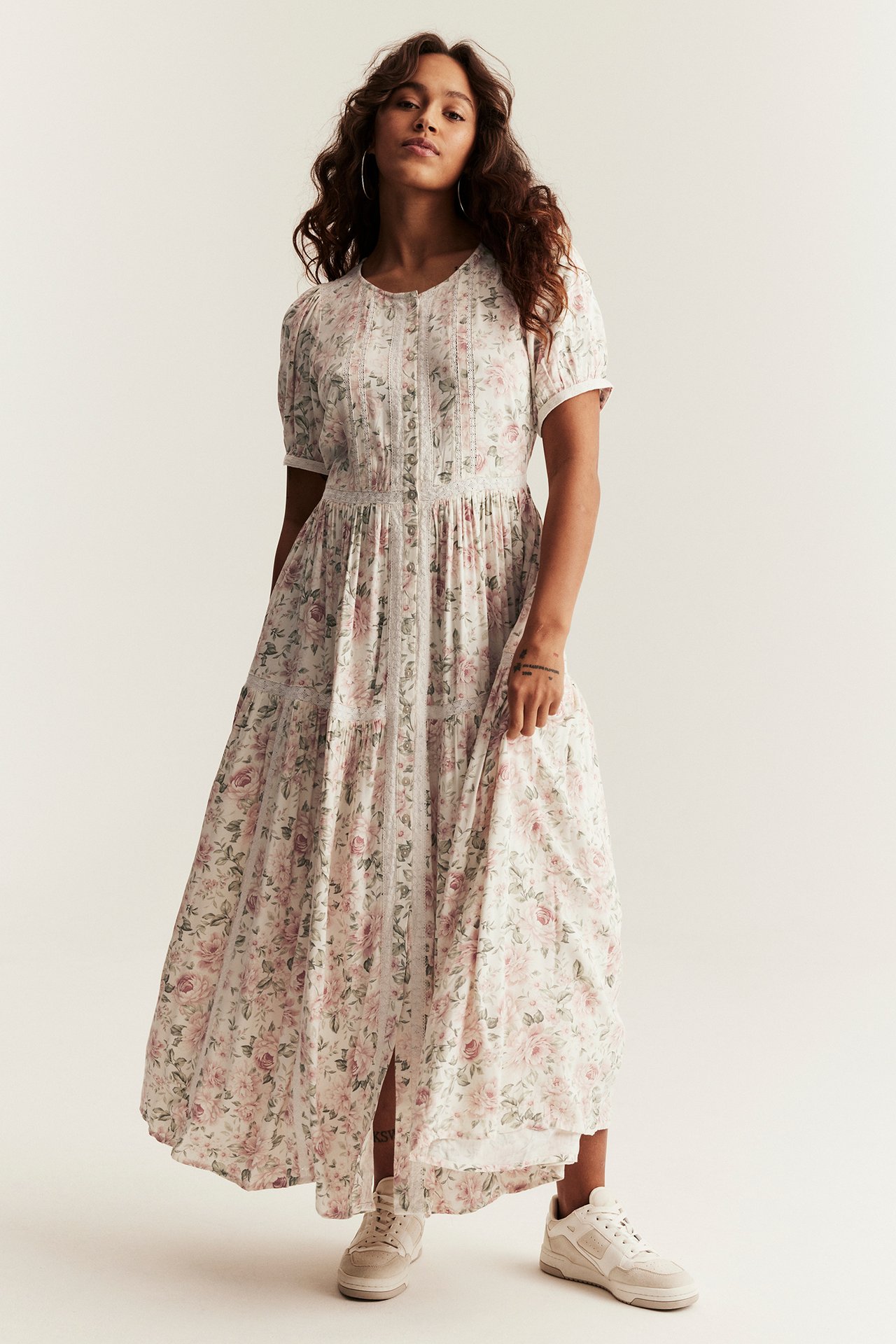 Vorderansicht: Frau trägt ein cremefarbenes geblümtes Maxikleid mit Puffärmeln, Spitze, Frontknöpfen und beigen Sneakers.