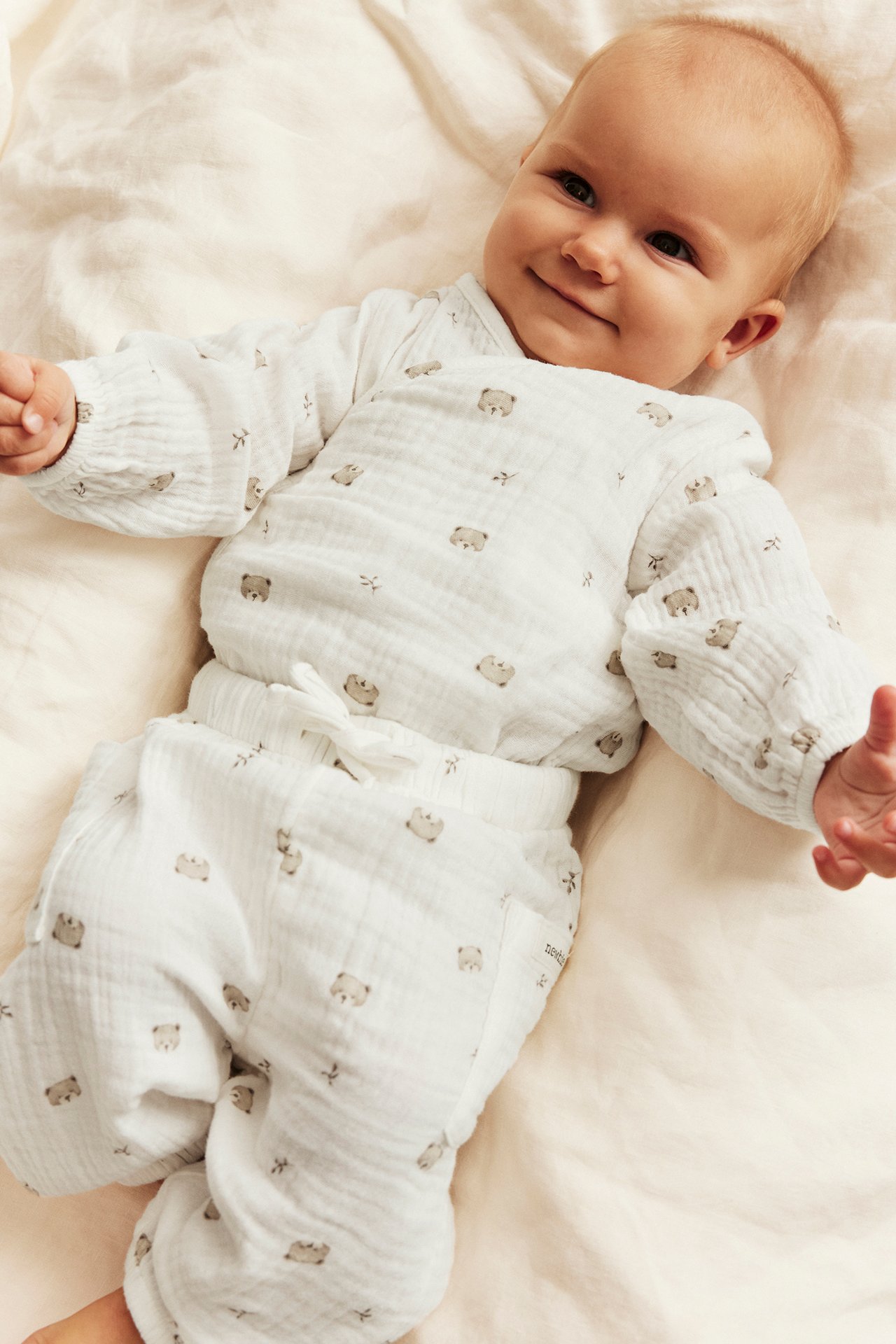 Vorderansicht von lächelndem Baby in weißem Musselin-Wickeloberteil und Hose mit Bärendruck.
