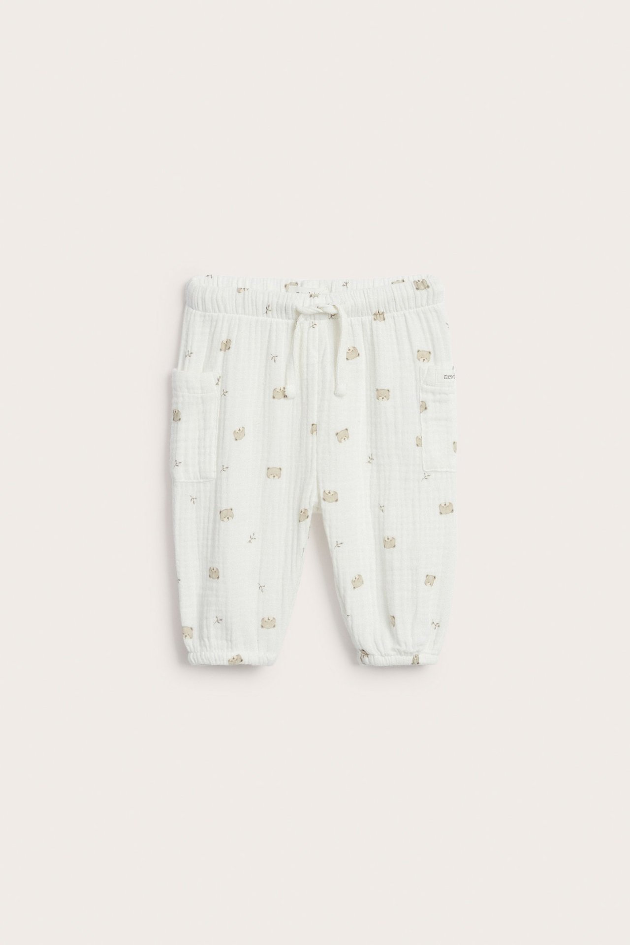 Front view: Off-white cotton muslin baby pants with bear print, elastic drawstring waist, and two patch pockets.
