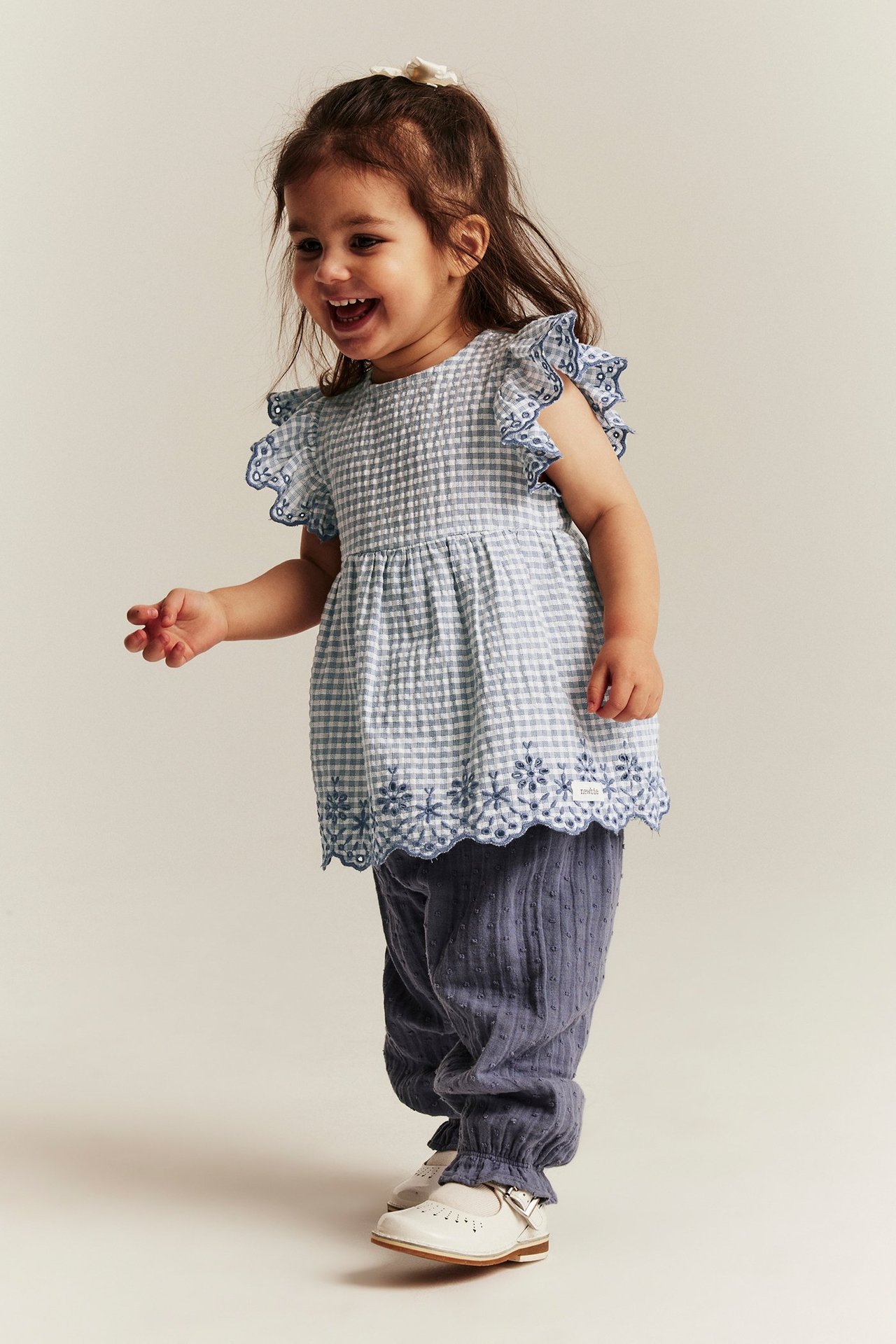 Vorderansicht: Ein Baby-Mädchen in blau-weiß kariertem Blusenbody mit Stickerei-Rüschen, blauen Hosen und weißen Schuhen.