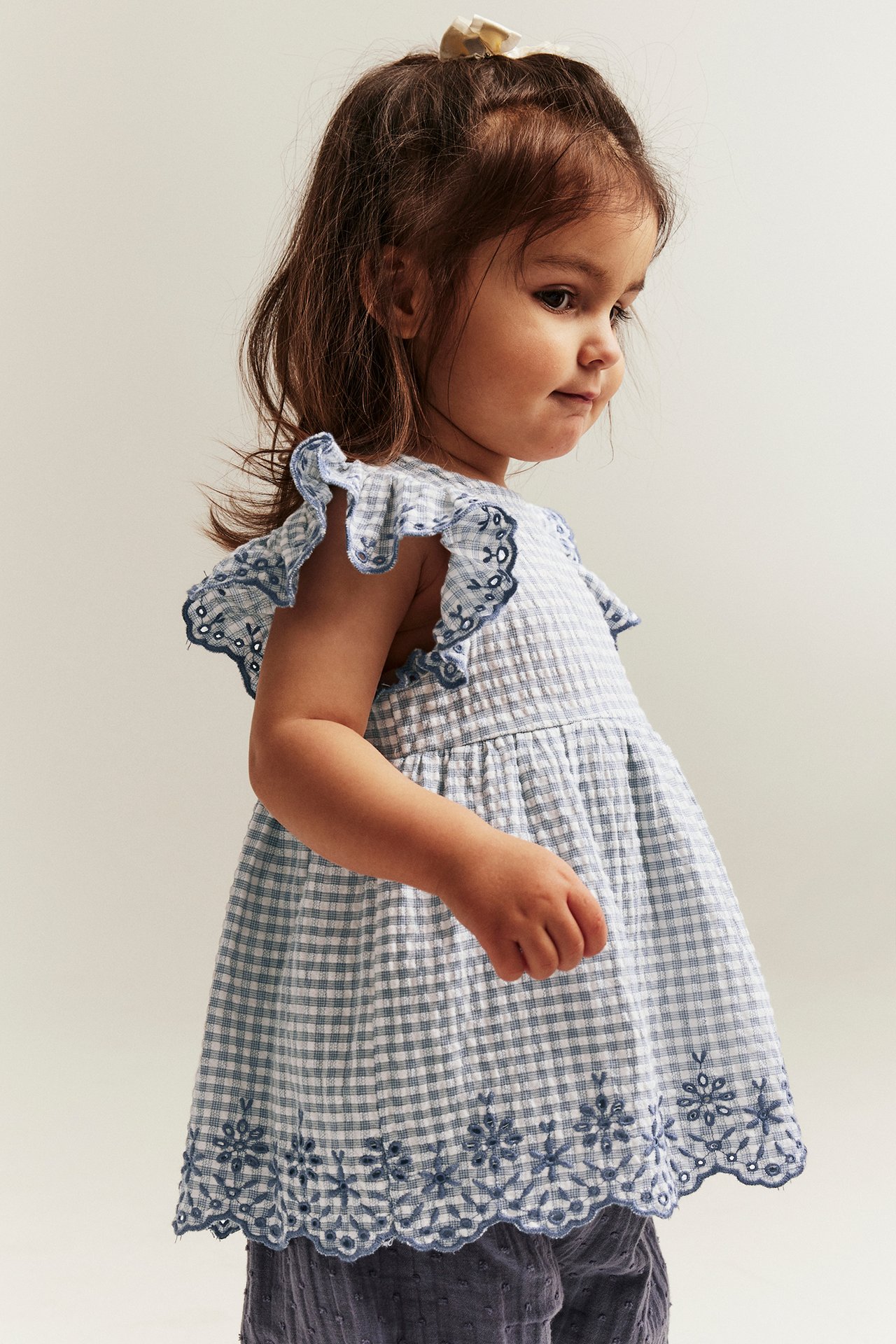 Side view of baby in blue and white checked cotton peplum bodysuit with embroidered ruffle sleeves.