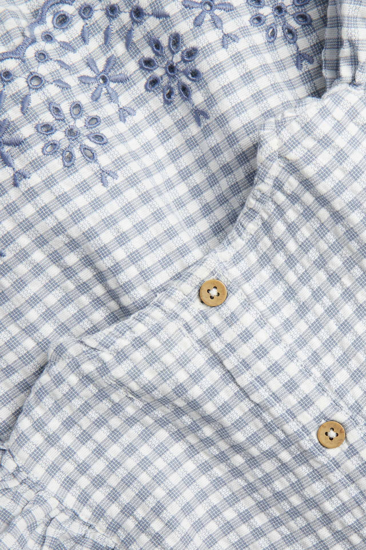 Nahaufnahme Baby-Blusenbody aus blau-weiß karierter Baumwolle mit Blumenstickerei und Holzknöpfen.