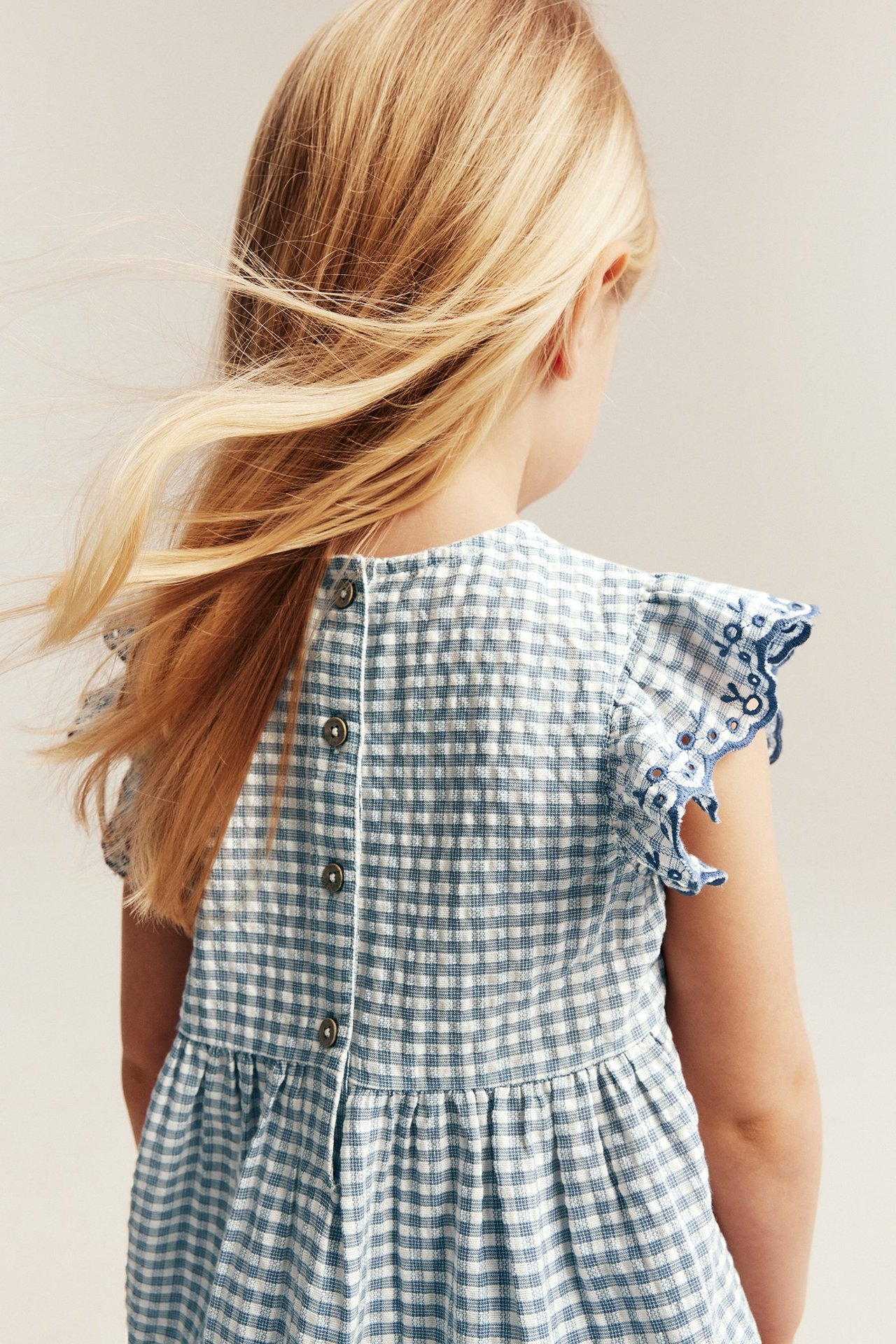 Back view of a kid wearing a light blue and white gingham cotton peplum blouse with embroidered ruffle sleeves and back buttons.