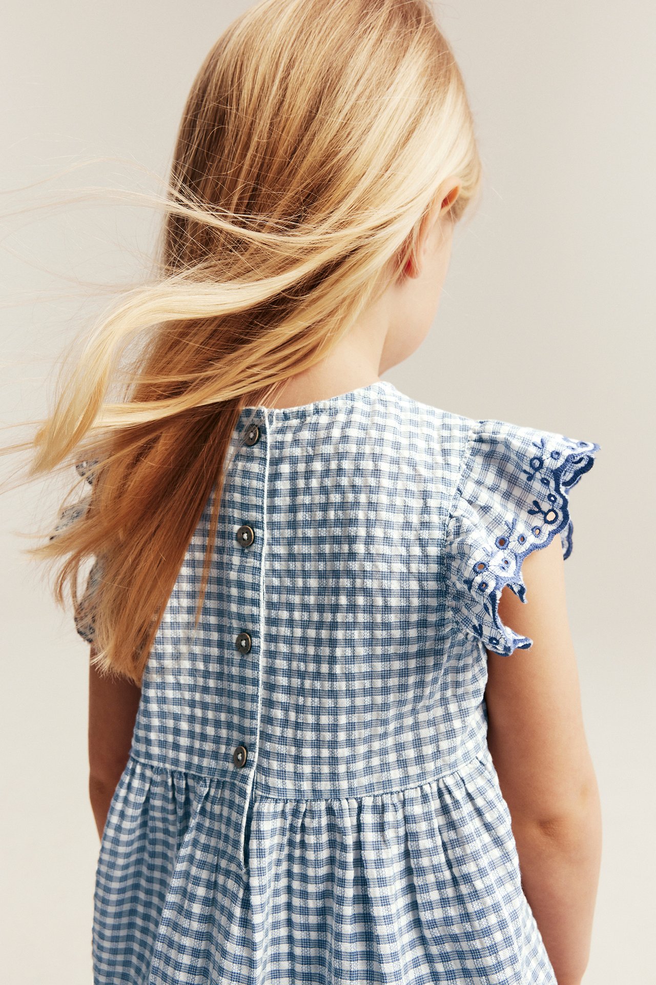 Back view of a kid in a blue and white checkered peplum blouse with embroidered ruffle sleeves and back buttons.