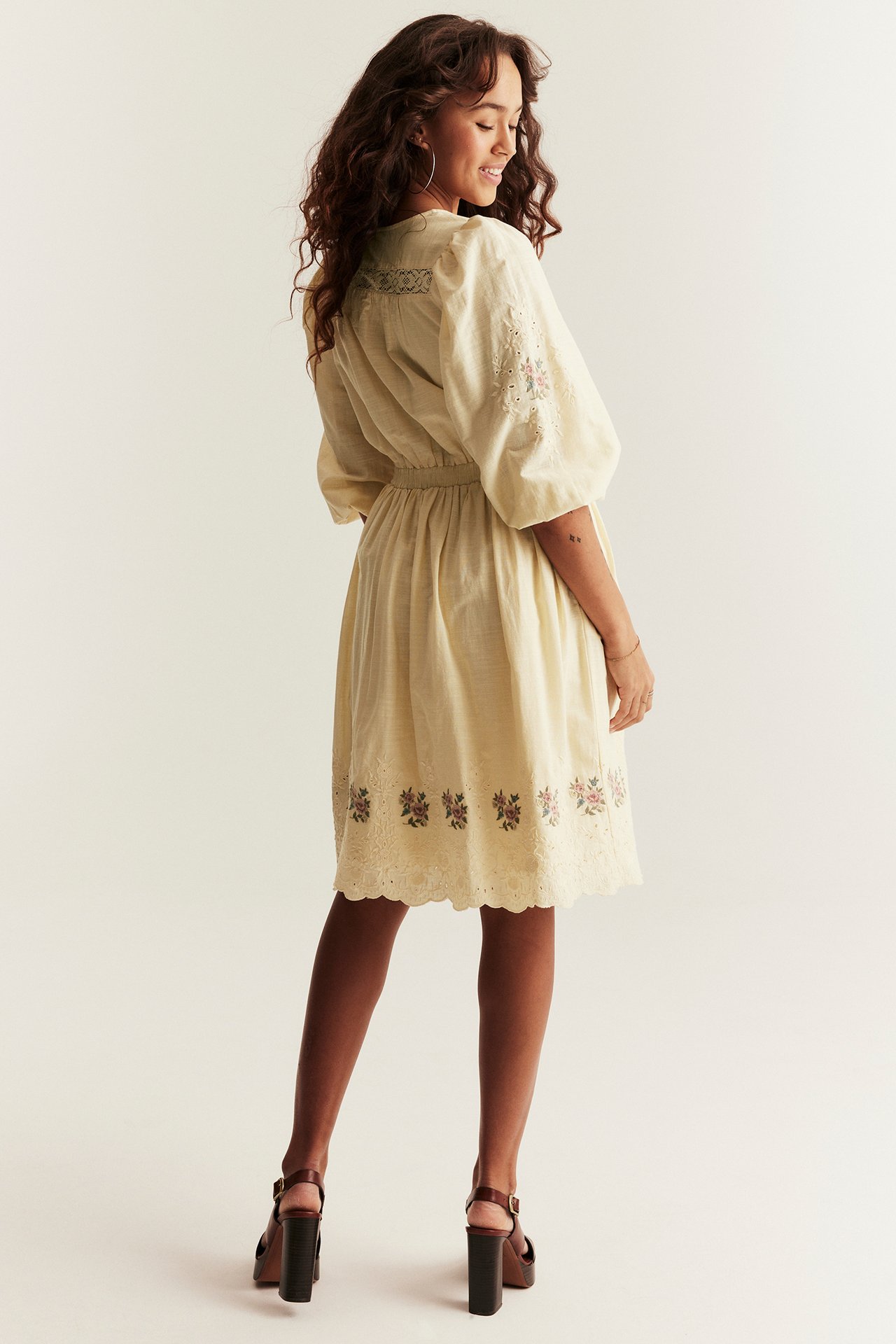 Back-side view, woman in a light yellow short dress with embroidery, puff sleeves, and lace, styled with brown sandals.