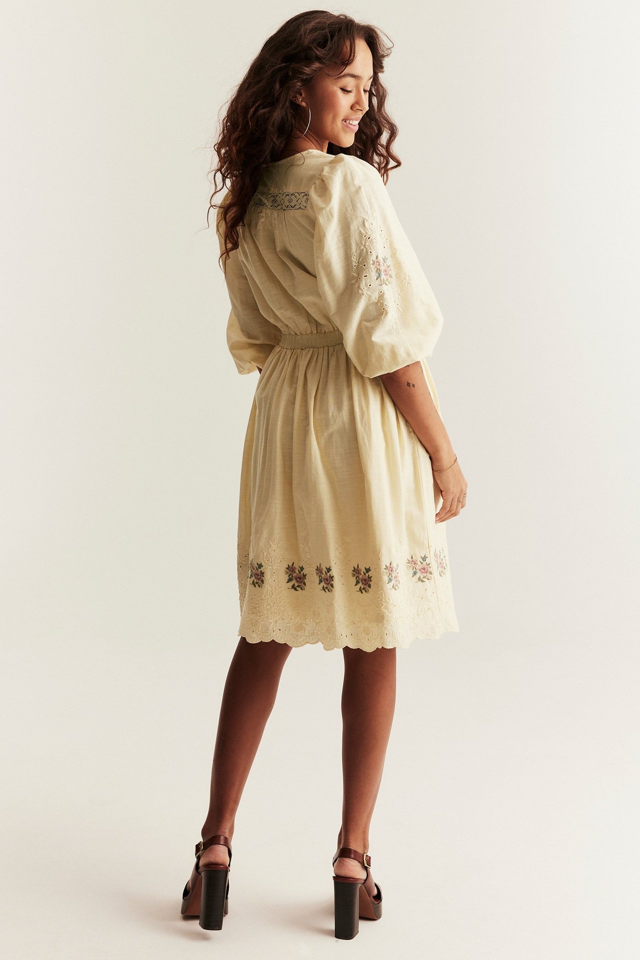 Back-side view, woman in a light yellow short dress with embroidery, puff sleeves, and lace, styled with brown sandals.