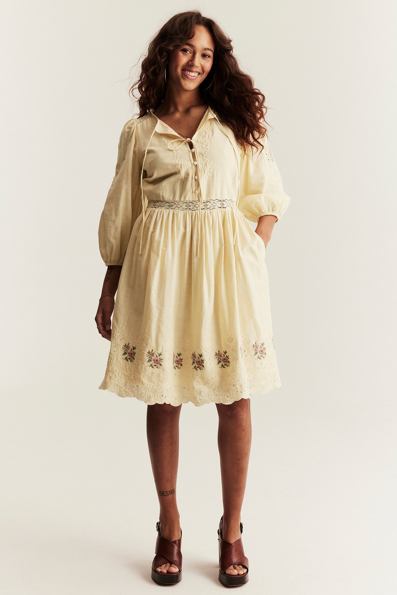 Vorderansicht einer lächelnden Frau in kurzem gelbem besticktem Kleid mit Puffärmeln und braunen Plateausandalen.