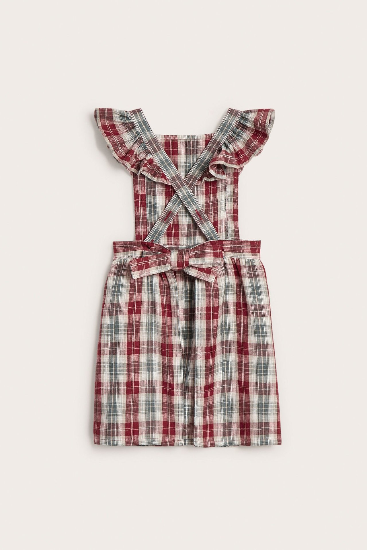 Front view of a kid's red, grey, and white plaid cotton apron dress with ruffled straps and a front bow.