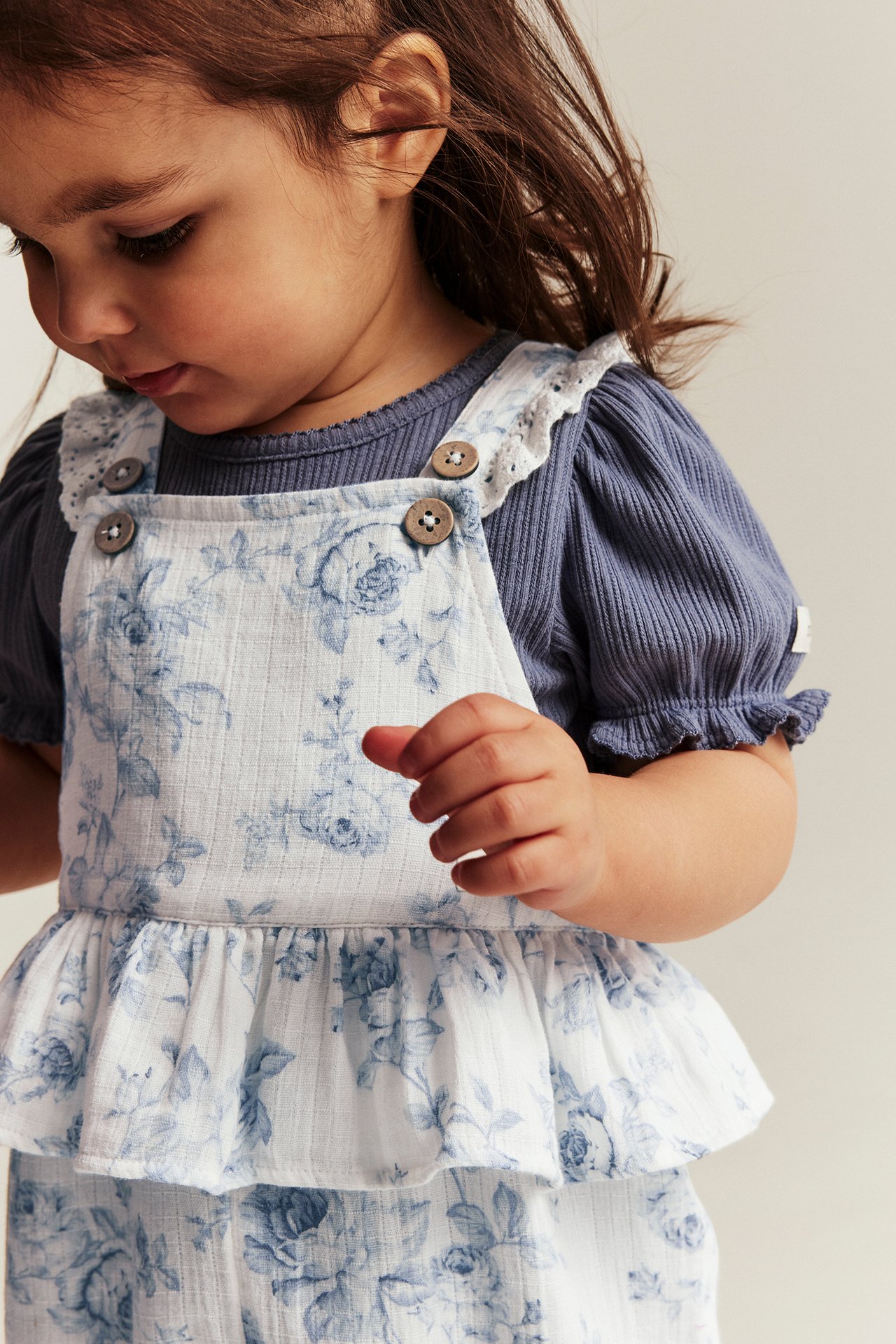 Close-up front view of a kid wearing off-white floral dungarees with lace trim over a blue ribbed top.